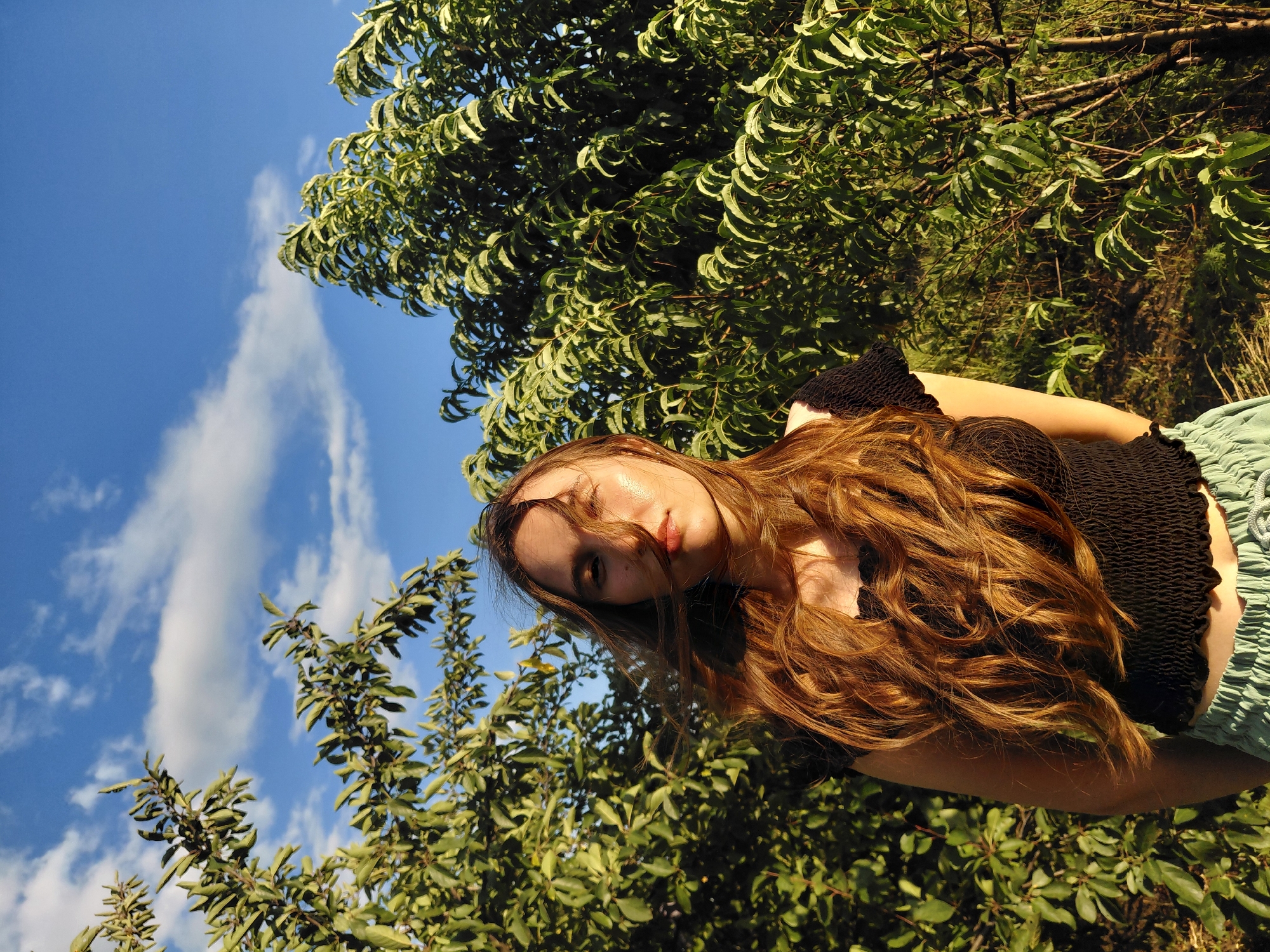 A young woman with long, wavy auburn hair is lying down, looking upwards towards the sky. Her face is partially obscured by her hair and the bright sunlight, but her eyes are visible. She is wearing a black ruffled top and light green bottoms. The background is filled with lush green foliage from trees and a bright blue sky with wispy white clouds. The scene appears to be outdoors, likely in a natural setting like a park or garden, during the daytime. The sunlight suggests it could be late afternoon or early evening. The overall mood is relaxed and serene. There is no visible text in the image.