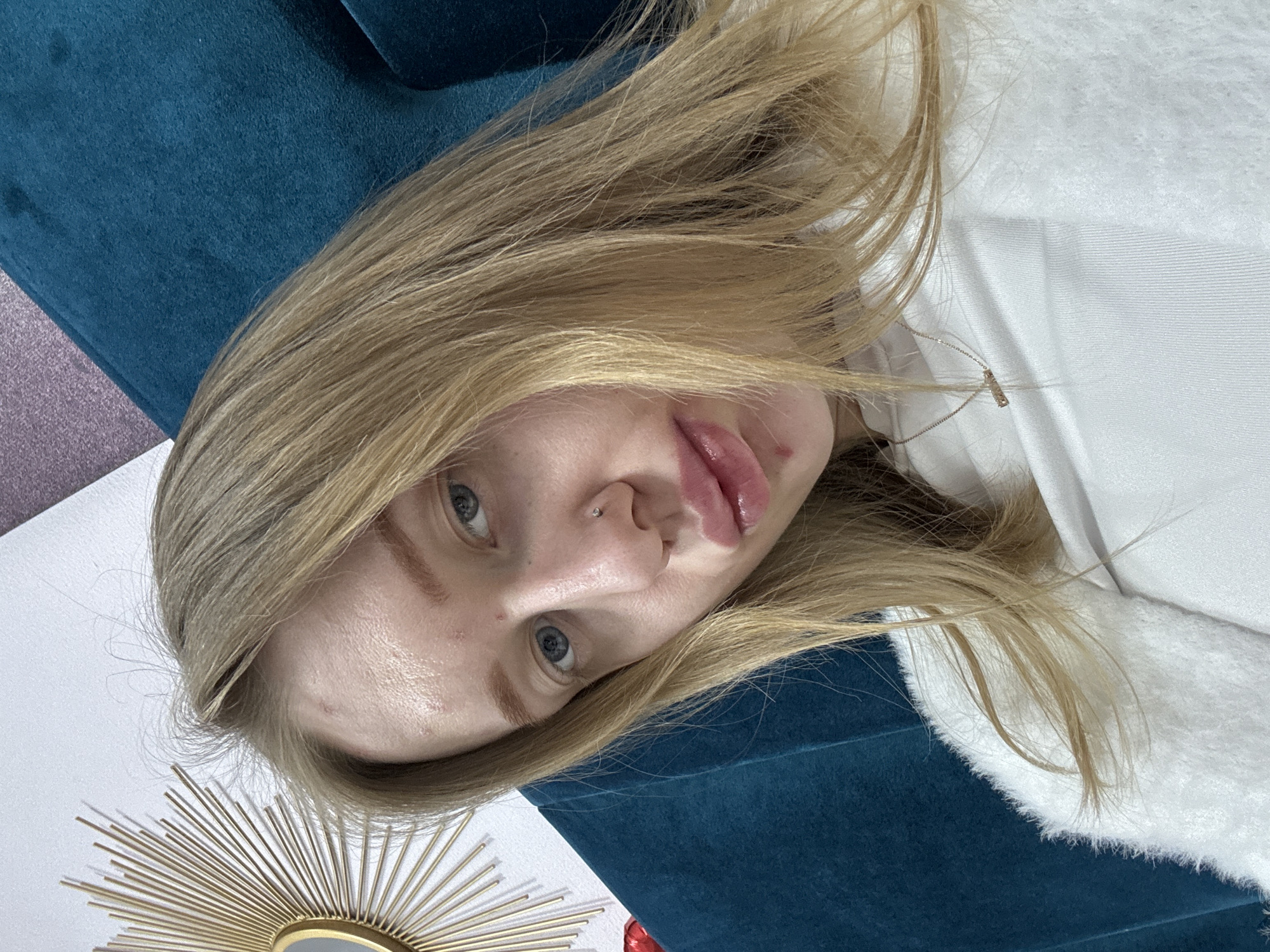 A portrait of a young woman with blonde hair and blue eyes. She has a nose piercing and is wearing a white garment. She is lying against a teal blue velvet surface. A golden sunburst-shaped decorative mirror is visible in the lower left corner. The location cannot be determined from the image. The city of Rostov-on-Don, Russia is not visually identifiable in the scene.