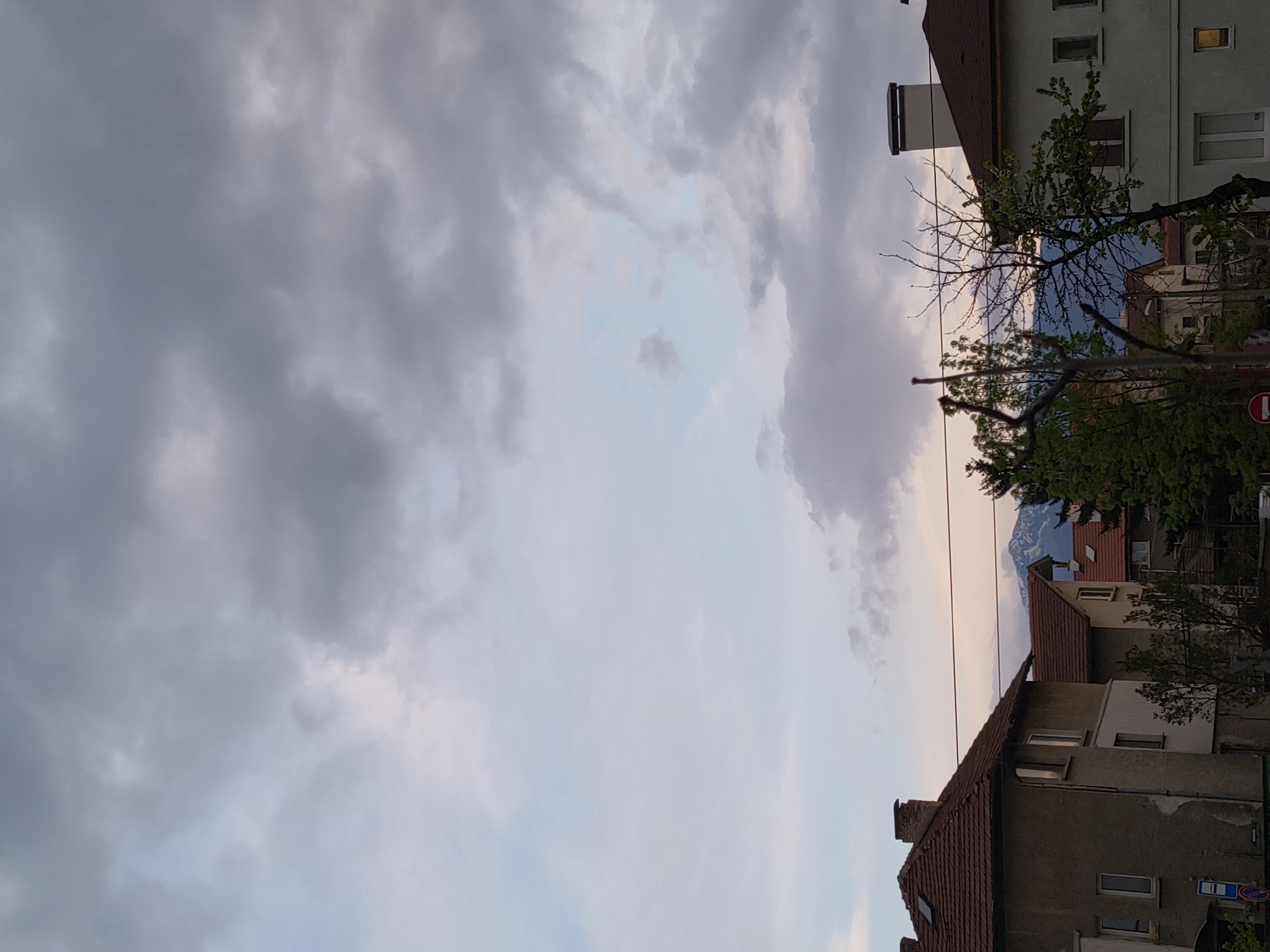 The image shows a view of Sofia, Bulgaria, facing south. The foreground consists of several buildings, mostly residential, with red tile roofs and light-colored walls. The trees in front of the buildings partially obscure the view. A mountain range with snow-capped peaks is visible in the distance between the buildings. The sky is mostly covered with grey and white clouds.