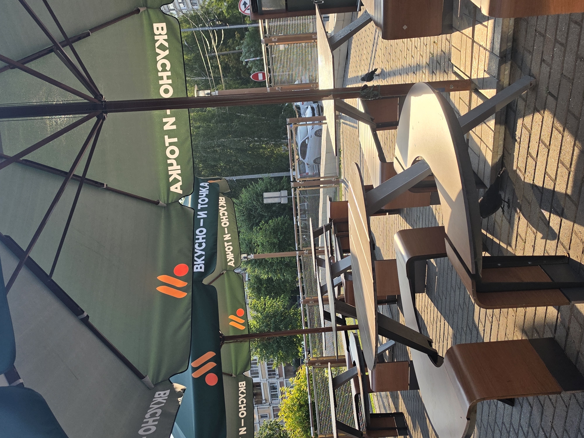 The image shows an outdoor seating area with picnic tables and umbrellas, likely at a fast-food restaurant. The umbrellas are green with the logo of "Vkusno i tochka" (Вкусно – и точка), a Russian fast-food chain that replaced McDonald's. Several picnic tables, made of wood and metal, are arranged on a brick-paved ground. Some tables have curved benches attached. Two crows are visible on the ground, pecking at something. In the background, there are trees, buildings, a road with cars, and streetlights. The scene appears to be bathed in the warm light of the setting sun, casting long shadows. The location is identified as Nizhny Novgorod, Russia.