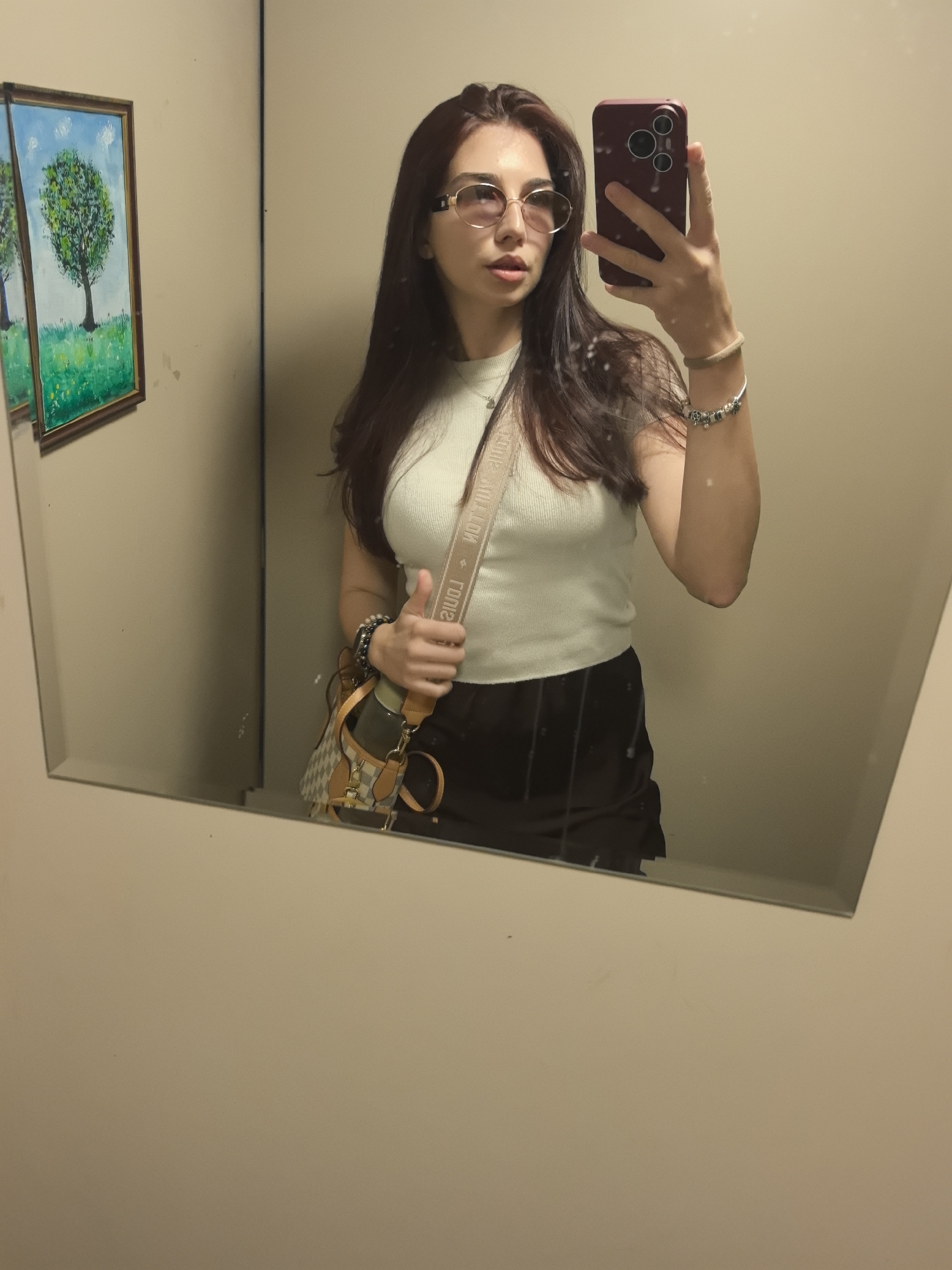 A woman is taking a selfie in front of a mirror. She has long, dark hair and is wearing sunglasses, a light-colored top, and a dark skirt. She is holding a phone in her right hand, and she has a purse hanging on her left shoulder. There are two framed pictures on the wall to the left of the mirror. The environment appears to be indoors, possibly a dressing room. There is no discernible information regarding the location being in Stara Zagora, Bulgaria.