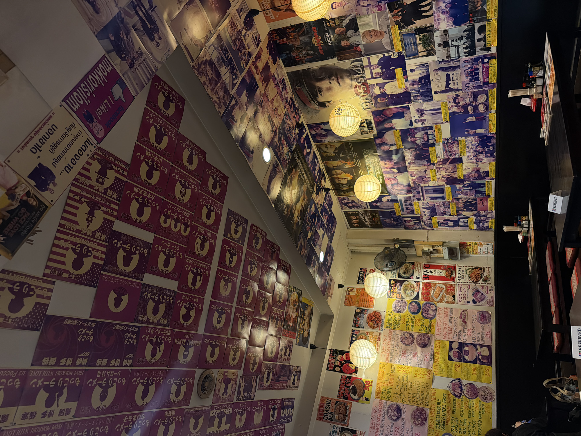 The image shows the interior of a Japanese ramen restaurant in Bangkok, Thailand, with the ceiling and walls heavily decorated with posters. The posters are a mix of Japanese text, silhouettes, movie posters, and portraits, creating a vibrant and eclectic atmosphere. Two large, white paper lanterns hang from the ceiling, providing ambient light. In the foreground, there are several dark wooden tables with red-cushioned benches, set up for dining. A portion of a table in the lower foreground has a "RESERVED" sign on it. On the walls, menus are displayed alongside more posters, showcasing various ramen dishes and their prices. A black electric fan is mounted on the wall. The overall impression is of a busy, themed restaurant.