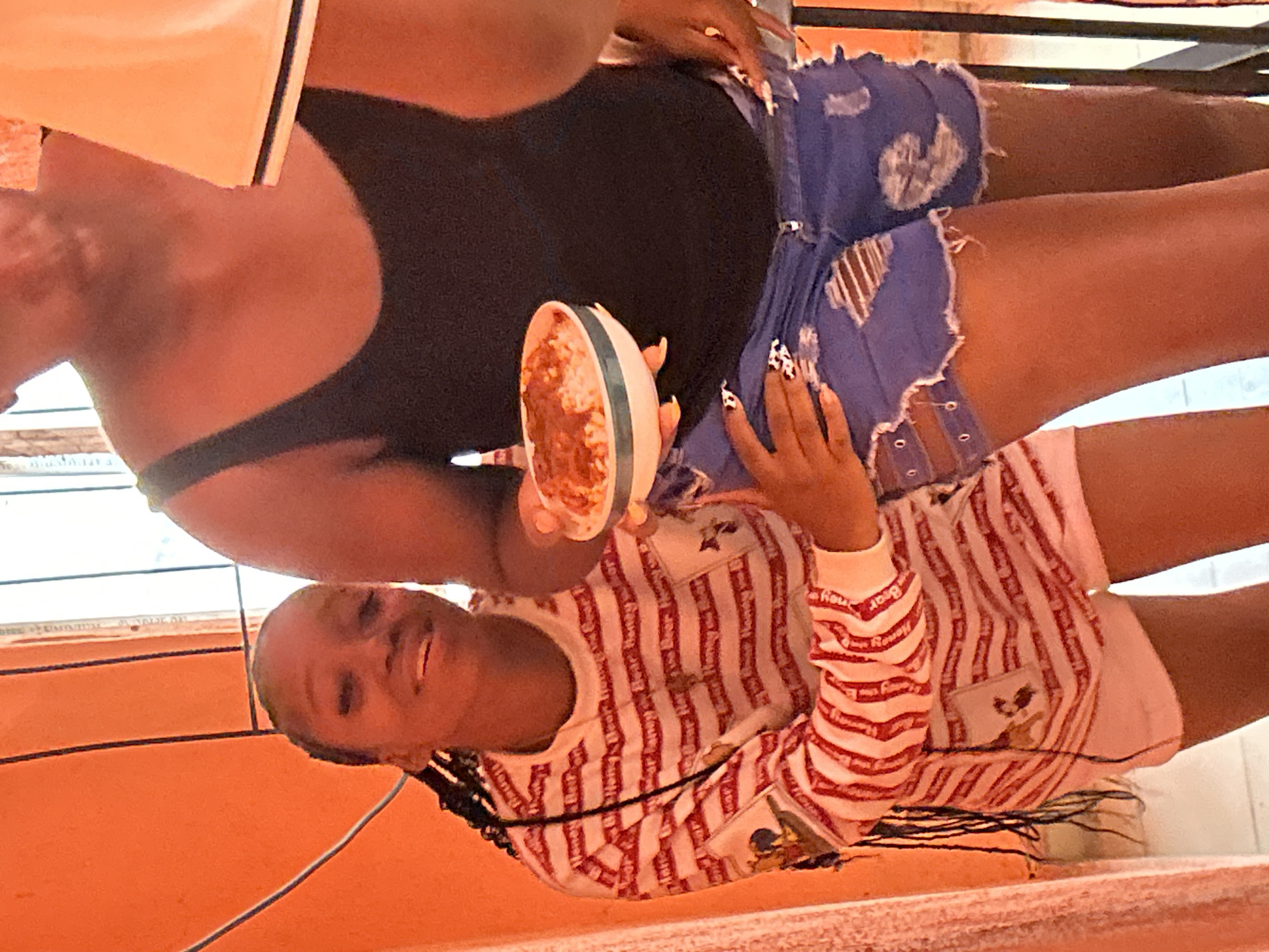 Two dark-skinned women are seated. One wears a black tank top and blue denim shorts with rips. She holds a bowl of food with a reddish sauce. The second woman wears a long-sleeved shirt with red and white stripes and cartoon graphics. The location appears to be indoors, possibly a residence in Ihiagwa, Nigeria, based on the visual features.