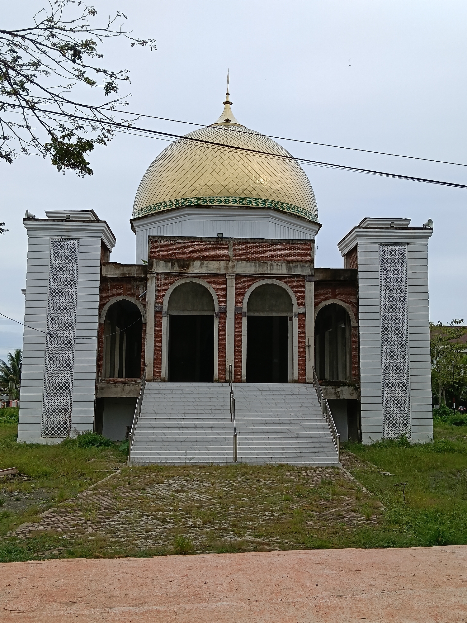 The image shows a mosque under construction in Meureudu, Indonesia. The mosque features a prominent golden dome with intricate green detailing around its base. The main structure is built with exposed red bricks and has large arched entrances. On either side of the entrance, there are tall, white pillars decorated with a repeating geometric pattern. A wide, white staircase leads up to the main entrance. The surrounding area is covered in grass and some unpaved ground, with a few trees visible in the background. The sky is overcast. There are no people visible in the image. The scene suggests ongoing construction work, with some elements of the building completed while others remain unfinished. The location appears to be a semi-urban or rural setting, given the vegetation and the general unfinished state of the grounds.