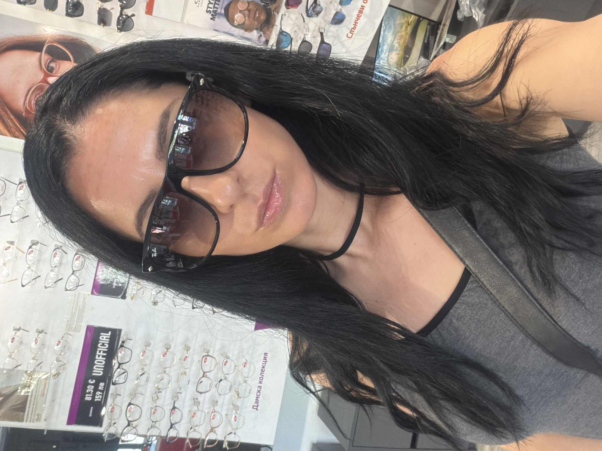 A woman with long, dark hair and large sunglasses is taking a selfie in what appears to be an optical store in Pleven, Bulgaria. She is wearing a grey tank top and a black choker necklace. In the background, there are displays of eyeglasses and sunglasses. A sign on the left reads "81.30 € UNOFFICIAL 159 лв". Another sign on the right has the text "STYLE MATTERS" and a picture of a man wearing glasses. The overall impression is a retail environment focused on eyewear.