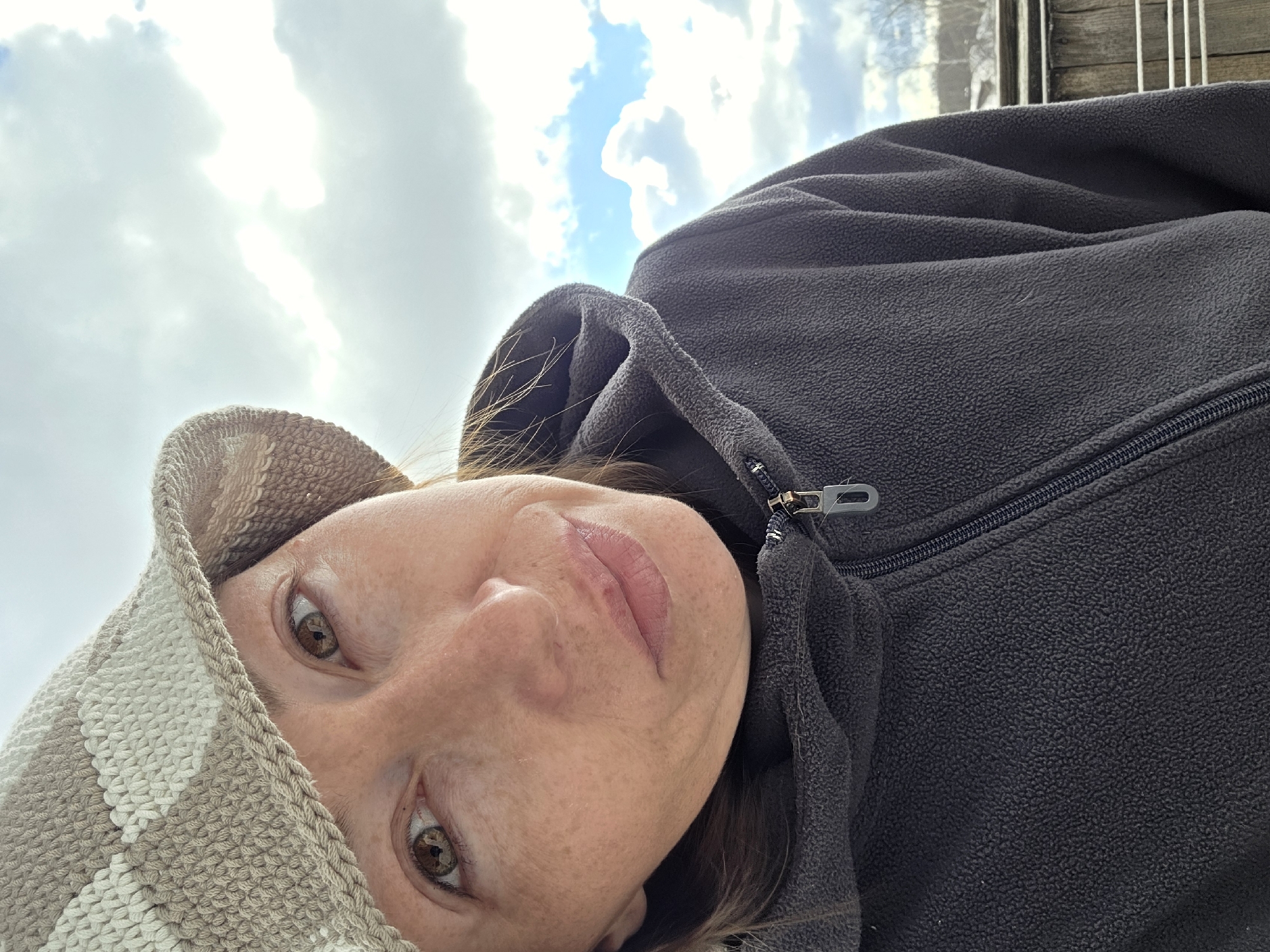 This close-up, diagonally oriented portrait features a person, likely a woman, looking upwards towards the camera or sky. She has light skin, brown eyes, and light brown hair partially visible around her face. Her expression is neutral and direct. She is wearing a dark grey or black fleece jacket, visible by its textured material and a silver-toned zipper and pull tab on the right side of the frame. Her head is partially covered by what appears to be a light brown and white striped or patterned knitted hood or hat, framing her face.
The background primarily consists of a bright, partly cloudy sky. Fluffy white clouds are scattered across the frame, with sections of clear blue sky visible between them. A very bright area in the clouds suggests the position of the sun. In the far upper right corner, a glimpse of a wooden structure, possibly a fence or railing, indicates an outdoor setting. The scene captures an outdoor moment under natural daylight, typical of Chaikovsky, Russia. There is no visible text in the image.