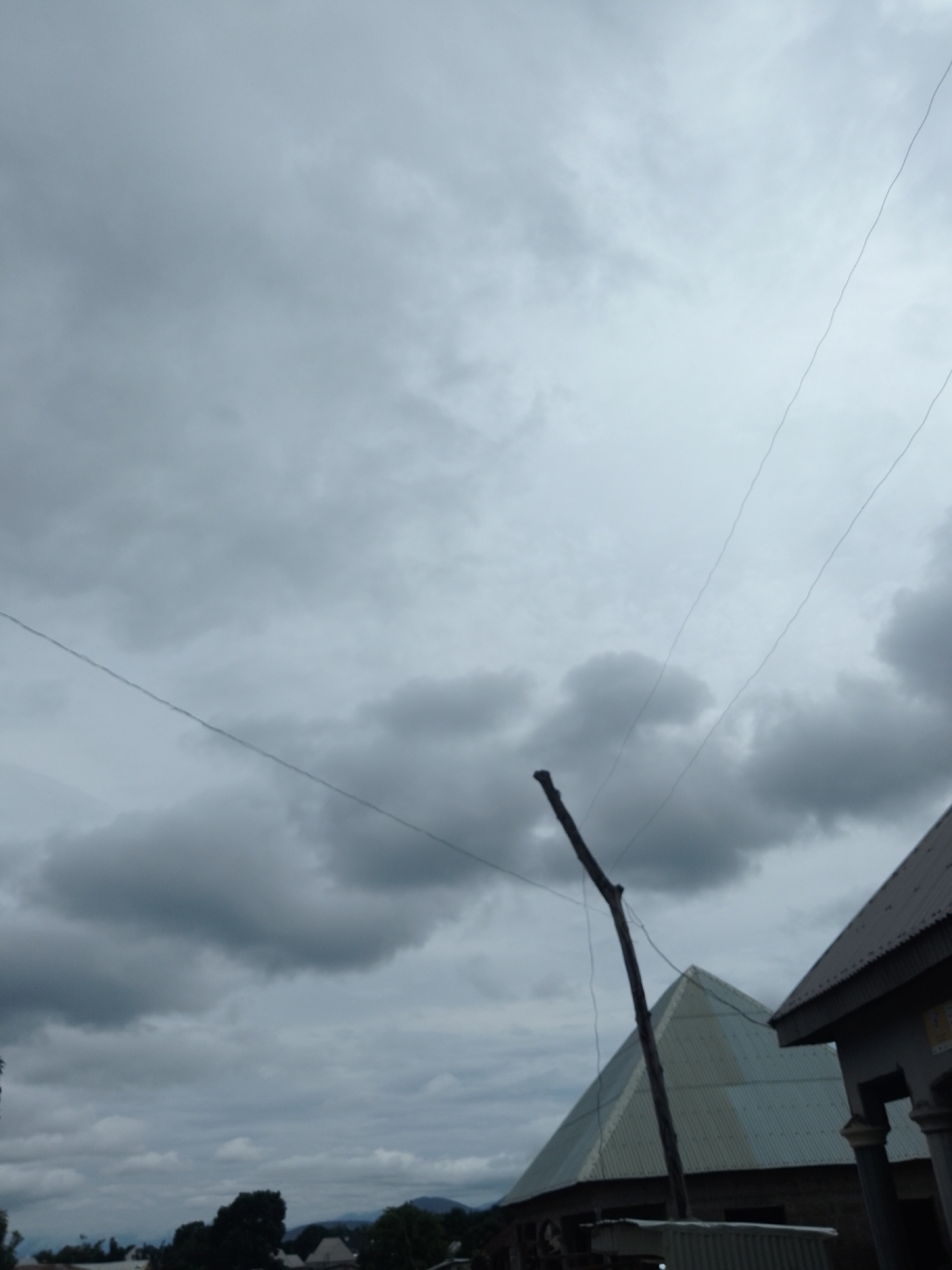 The image captures an overcast sky above a cluster of buildings in Serti, Nigeria. The sky is filled with dark, heavy clouds, suggesting an impending rain or a storm. In the foreground, a wooden pole stands vertically, with wires attached to it. To the right, the corner of a building with a grey, corrugated metal roof is visible, featuring pillars and a sign. Further to the left, another building with a pyramid-shaped, light-colored corrugated metal roof is partially visible. The lower portion of the image reveals a dark, silhouetted landscape with trees and more rooftops, indicating a populated area within the city. The overall scene is somber due to the dark clouds and limited visibility.