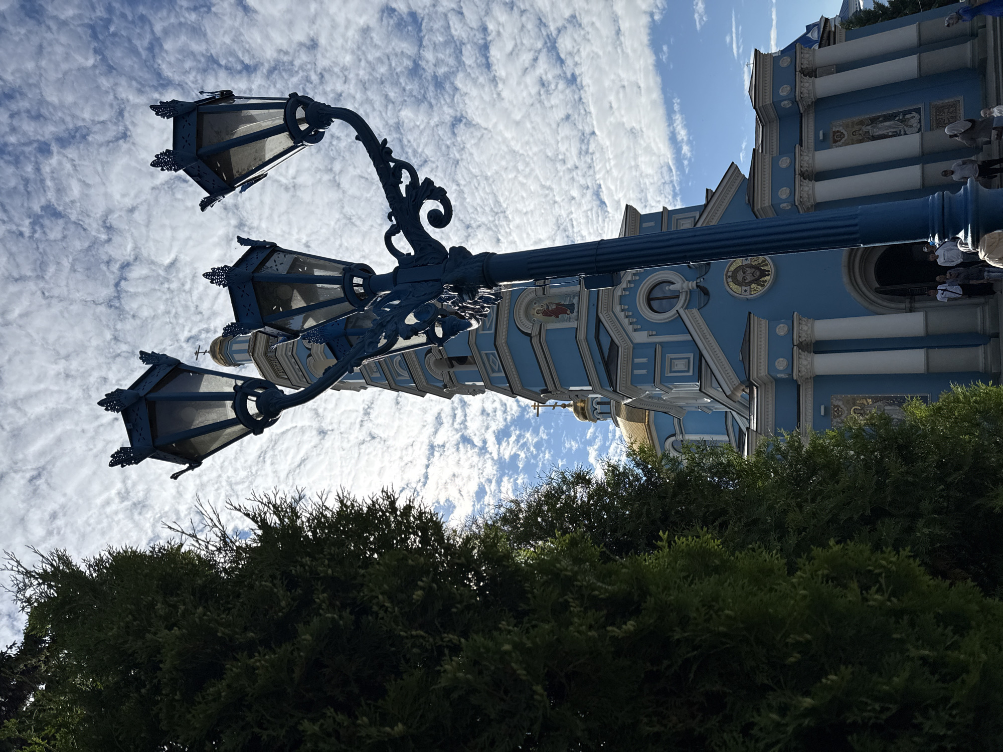 An outdoor, low-angle, medium shot captures a bright blue Orthodox church in the city of Unknown. The church features golden domes, white columns, and mosaic religious imagery. In the foreground, a large, ornate, dark blue lamppost with three lamps stands to the left, its decorative elements partially obscuring the view of the church and a dark green evergreen tree to the left of the lamppost. The sky above is a vibrant blue with scattered white, fluffy clouds. Several people are visible near the entrance of the church, some standing and looking at the building, while others appear to be walking or posing for photos.