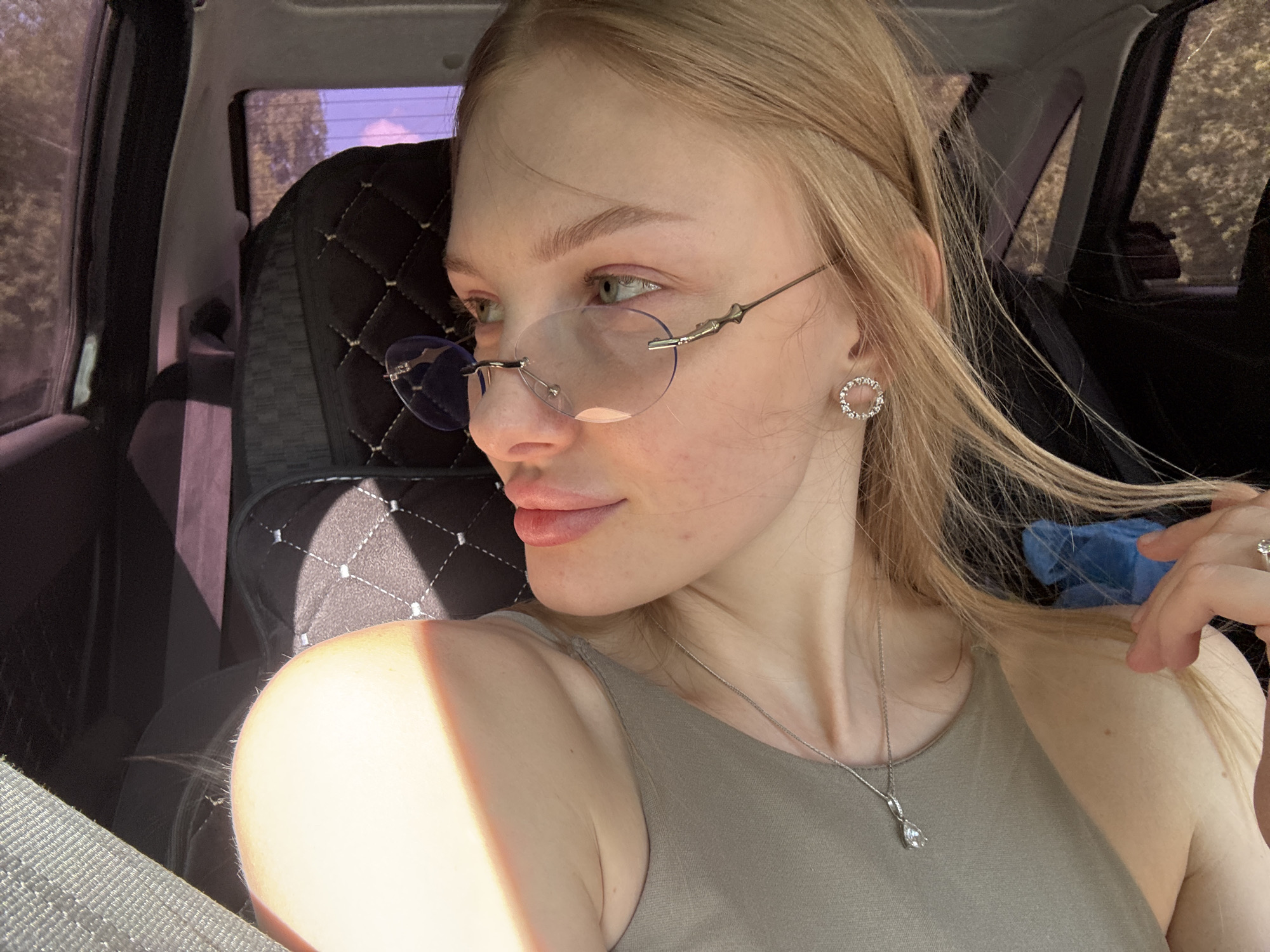 A young woman is sitting in the driver's seat of a car. She is fair-skinned with blonde hair and is wearing glasses. She also has an earring and a necklace. The interior of the car is visible, including the seat cover and window. The location is Perm, Russia.