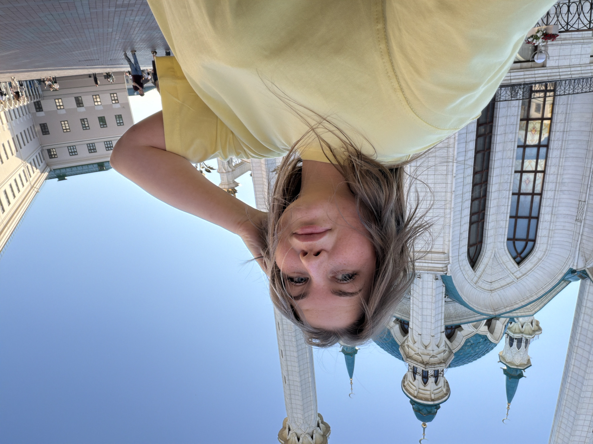 A young woman, appearing to be in her 20s, is centered in the foreground, capturing a selfie from a low angle. She has light brown hair, blue eyes, and freckles on her nose and cheeks. She wears a loose, light yellow t-shirt, and her left hand is raised, with fingers touching the back of her head. Her lips are slightly parted. The primary background feature is the Kul Sharif Mosque, situated in the Kazan Kremlin, Kazan, Russia. The mosque exhibits prominent white stone architecture with arched windows, a large blue-green tiled dome, and multiple minarets, all topped with golden crescent moons. To the right of the woman, a paved public square is visible, occupied by several small figures walking or standing in the distance. A large, light-colored building with numerous rectangular windows lines the right side of the square. The sky is clear and light blue, indicating a bright, possibly late afternoon or early evening, on a sunny day.