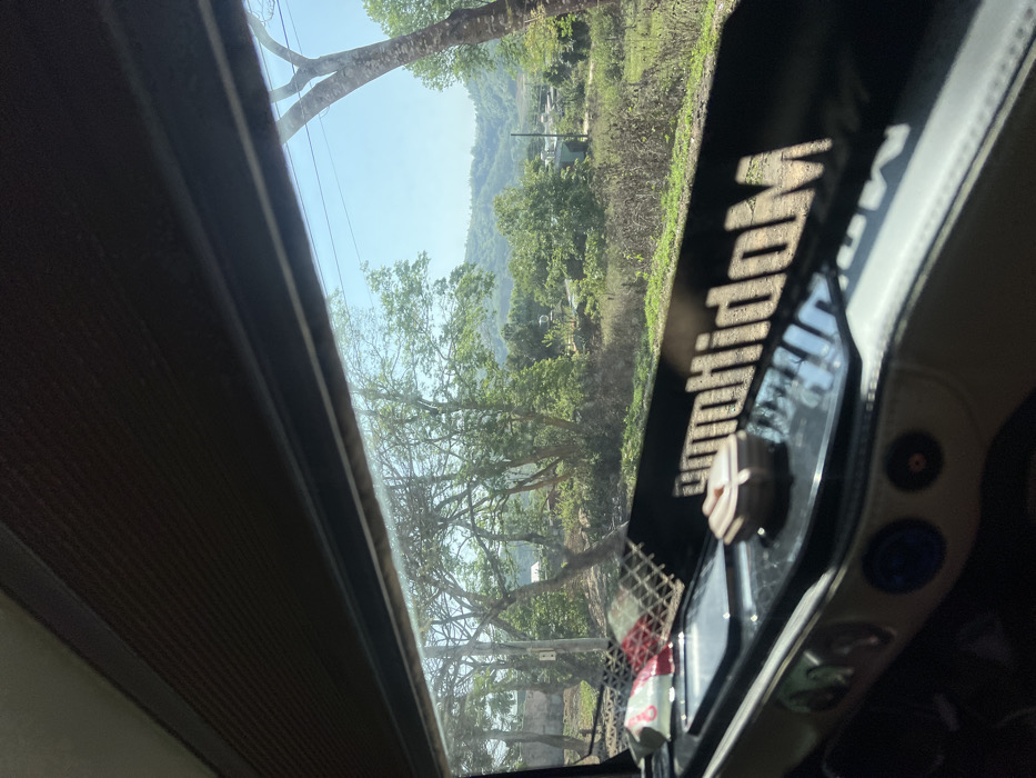 The image captures a view from inside a vehicle, looking out a side window onto a rural landscape in Pờ Y, Vietnam, on a clear, sunny day. The top of the frame shows a dark, textured overhead interior panel. A dark window frame separates the vehicle's interior from the exterior scene.
In the immediate foreground, part of the vehicle's dashboard is visible. It features a black, glossy surface with a reflective white inscription that reads "MudihdoM" (likely "Modihdum" or similar, mirrored). On this surface, from left to right, are a white object with a red pattern (resembling a tissue packet or snack wrapper) held within a mesh compartment, a flat rectangular object (possibly a smartphone), and a small, light-brown, multi-faceted container. Below this black surface, a beige/white padded section of the dashboard with circular control elements or vents is partially visible.
Outside the window, a vibrant green landscape unfolds under a clear blue sky. Numerous trees, some with dense foliage and others appearing sparser, dominate the midground. A utility pole with overhead wires is visible amidst the trees on the left side. Further back, scattered small structures or houses are discernible among the vegetation, suggesting a rural or semi-rural settlement. The background features rolling green hills under the bright sunlight. No people or animals are visible in the scene.
