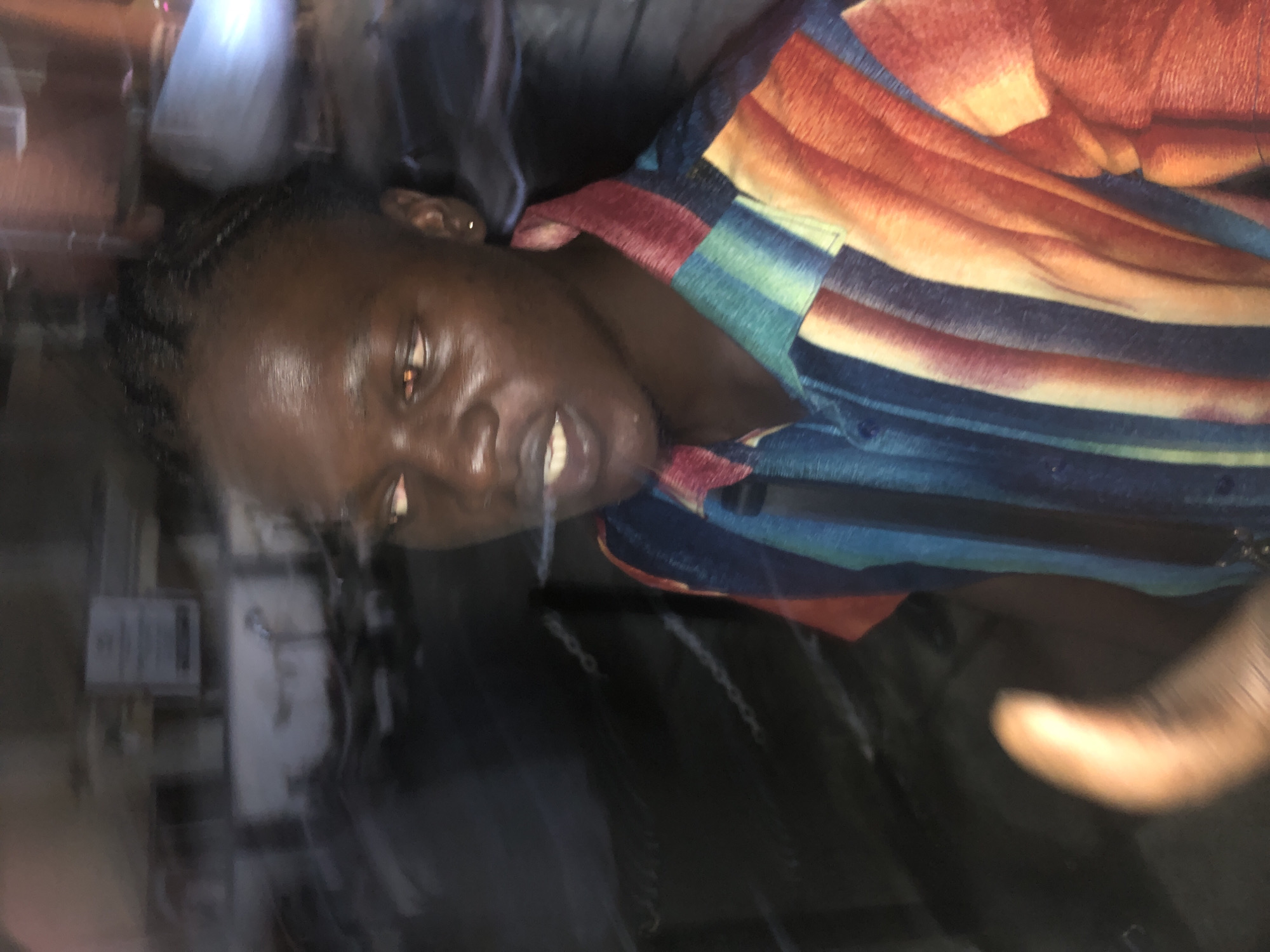 The image shows a blurred close-up of a dark-skinned man from the chest up, facing slightly to the right. He has braided hair and is wearing a brightly colored, vertically striped shirt with hues of blue, green, orange, and red. His eyes are open and appear to have a reddish-orange glow. His mouth is slightly open, and it appears he might be speaking or exhaling. A hand, blurred and partially in focus, is visible in the lower left corner, with its thumb pointing upwards. The background is dark and heavily blurred, with some indistinct shapes that suggest an outdoor urban environment at night. The overall impression is one of motion blur, possibly due to a moving camera or subject. It is not possible to determine the specific location in Ado, Nigeria from the visual information alone.