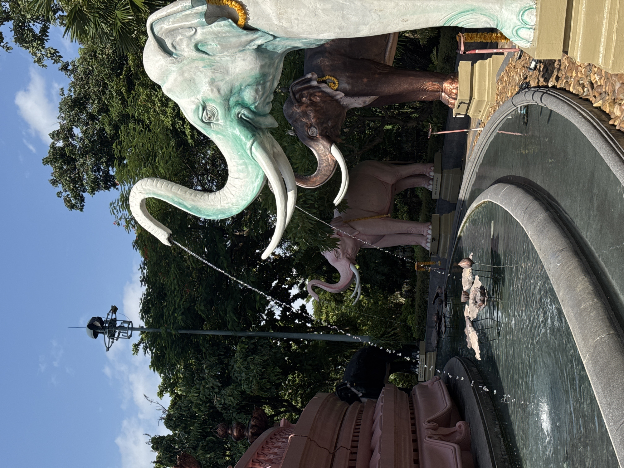 In Samrong Nuea Subdistrict Municipality, Thailand, a sunny day features a decorative fountain area. The foreground shows a curved, dark gray basin filled with dark, rippling water, with several metal lotus flower sculptures emerging from the surface. Water cascades from multiple sources, including a large, light green elephant sculpture whose trunk sprays water into the basin. Behind it, a dark brown elephant sculpture stands, with a pink elephant sculpture further in the background. To the left, a large, tiered, pink structure with decorative carvings is visible. In the mid-ground, a tall pole with a light fixture and what appears to be a speaker rises against a bright blue sky dotted with white clouds. Lush green trees form a dense canopy behind the elephant sculptures and the fountain. The ground surrounding the fountain is covered with small, light brown stones.