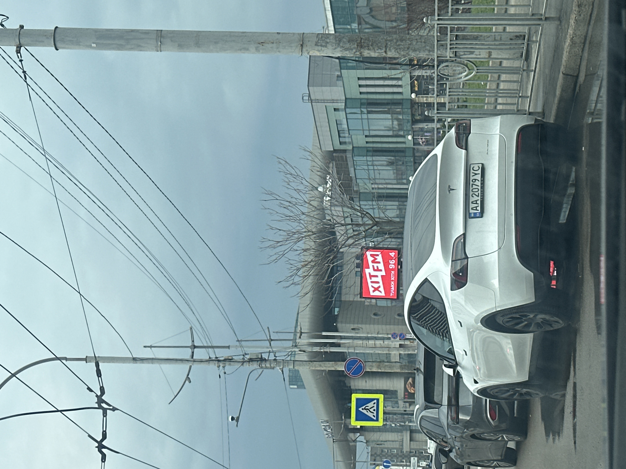 The image captures a street scene in Kyiv, Ukraine, on an overcast day. A white Tesla with the license plate "AA 2079 YC" is prominently featured, stopped in traffic. In the background, street signs, including a pedestrian crossing and "no stopping" sign, are visible, along with a large billboard advertising "XITFM" radio station at 96.4 FM. Power lines and utility poles line the street, and a modern building is partially visible in the background. Other vehicles are in the traffic lane beside the Tesla. The image is taken from inside another vehicle.