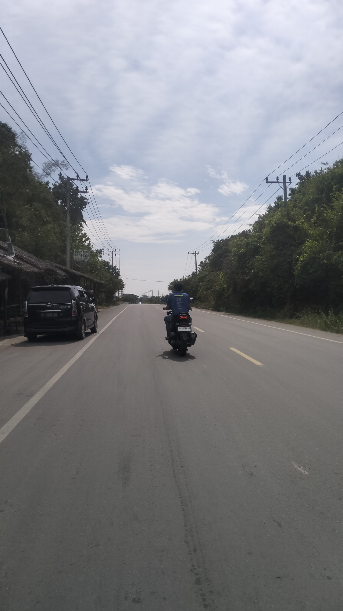 The image shows a wide, paved road in Lhokseumawe, Indonesia, with a slight upward slope. On the left side of the road, a dark-colored van is parked next to a small, open-air structure with a thatched roof. The van has a license plate that appears to be Indonesian. In the center of the road, a person is riding a motorcycle away from the camera. The motorcycle has a license plate. The rider is wearing a blue shirt with white lettering. The road is flanked by lush green trees on both sides. Overhead, power lines are strung between utility poles. The sky is partly cloudy. The scene suggests a rural or semi-rural setting on a clear day.