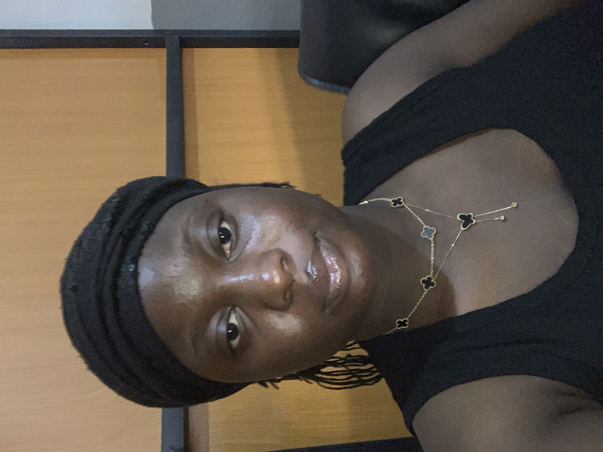 A medium shot captures a young woman from the chest up, seated indoors. She is wearing a black headscarf and a black tank top, adorned with a gold necklace featuring clover and butterfly charms. Her skin is dark, and her eyes are brown and directed towards the camera. The background consists of a wooden wall and a portion of a dark chair. The lighting is soft, creating a warm glow on her face. The scene appears to be a casual, personal moment.