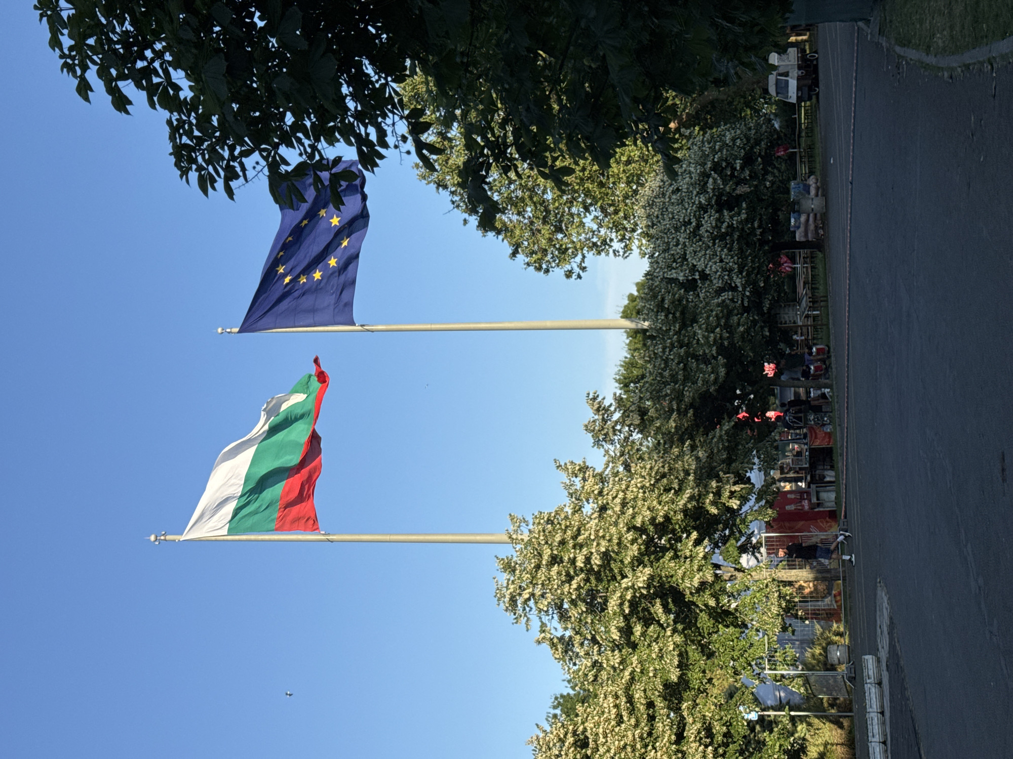 In Burgas, Bulgaria, two national flags, the Bulgarian flag and the European Union flag, are prominently displayed on tall flagpoles against a clear blue sky. The Bulgarian flag, a horizontal tricolor of white, green, and red, is positioned to the left and waves slightly. To its right, the EU flag, a blue rectangle with a circle of twelve yellow stars, is also airborne. Both flags are framed by the lush greenery of trees, with some trees in the foreground appearing to be in bloom with white flowers, and darker green foliage on the right.
Below the flags, a paved area suggests an outdoor public space. In the mid-ground, several trees provide shade over a scene that includes a white tent structure with metal fencing around it. Behind the fencing, there appear to be stalls or booths, possibly for a market or event, with some people visible. There are picnic tables and benches set up, suggesting an area for gathering or relaxation. A small, three-wheeled vehicle, a Piaggio Ape, is parked to the right, near what looks like more tents or structures. A few individuals are present in the scene, including one person walking on the paved area in the mid-ground. The ground is a dark asphalt surface, with a curb on the right side. The overall impression is of a pleasant outdoor setting on a clear day, possibly a park or a public square hosting an event.