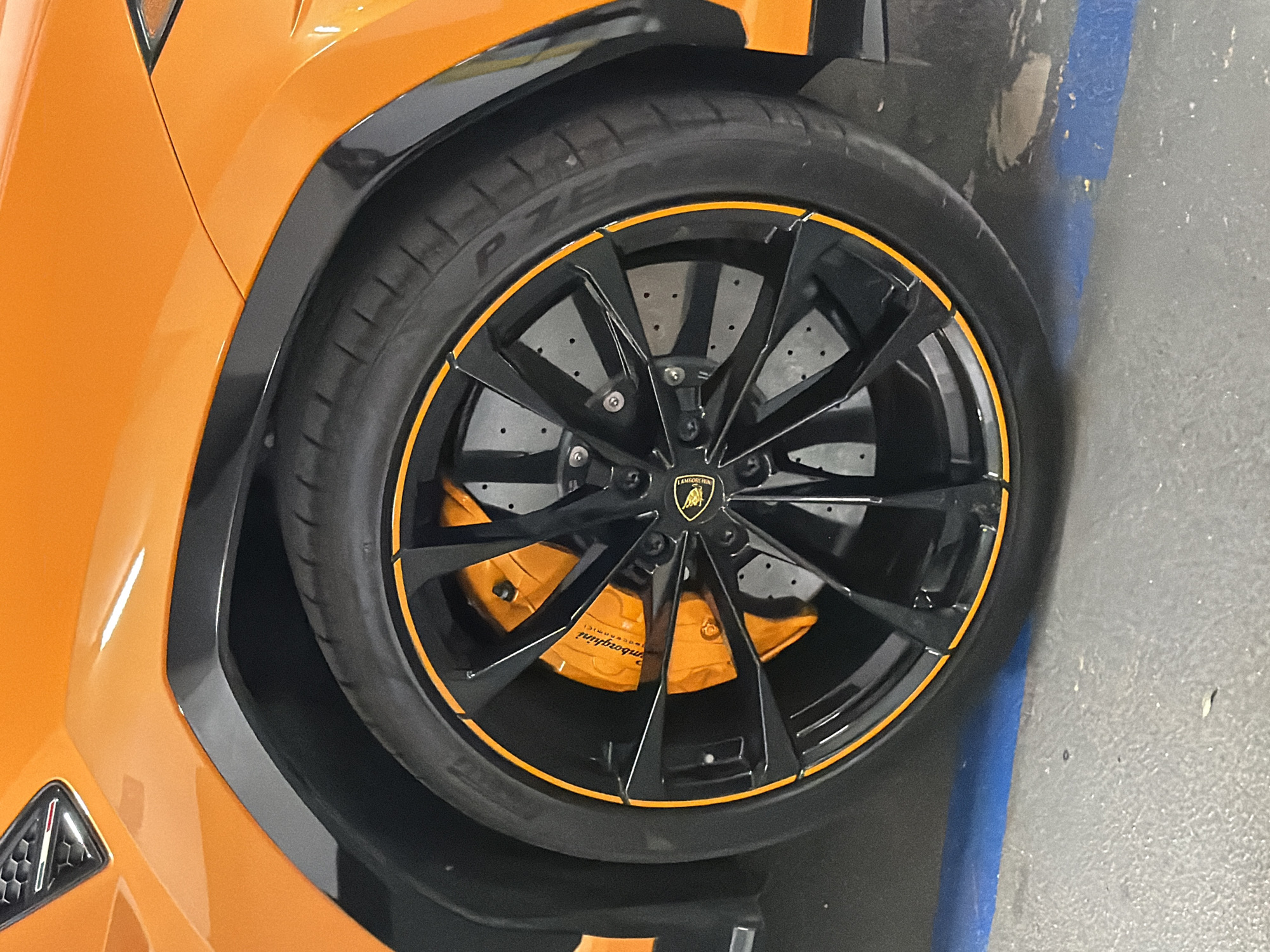 A close-up, eye-level, interior shot captures the front driver's side wheel and fender of an orange Lamborghini Urus. The car is parked in what appears to be an indoor parking garage, as indicated by the concrete floor and a faint blue painted line on the ground. The wheel is black with a bright orange accent on the rim. The brake caliper is also bright orange and prominently displays the Lamborghini logo. The car's bodywork is a vibrant orange, and a black fender flare arches over the tire. The image is well-lit, with sharp focus on the car's details. The background is blurred, drawing all attention to the vehicle.