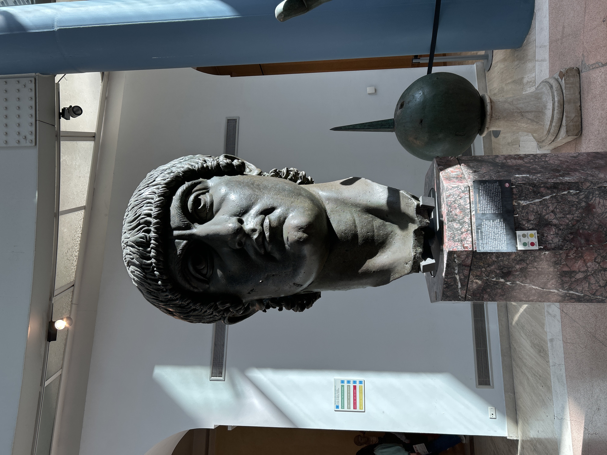 The image displays the colossal bronze bust of Constantine, situated within the Capitoline Museums in Rome, Italy. The prominent bust, depicting a male head with short, textured hair and a stern countenance, rests on a polished dark reddish-brown marble pedestal. An informational plaque with text and multi-colored indicators is attached to the pedestal. To the right, a dark green spherical artifact, topped with a conical spike, is presented on a light-colored, fluted stone base.
The setting is an indoor museum gallery characterized by white walls. Natural light filters in from overhead translucent panels, casting distinct shadows, while artificial spotlights on a ceiling track provide additional illumination. A robust metal beam with visible rivets crosses the upper frame, and a large, light blue cylindrical column is prominent on the far right. Horizontal air vents are visible on the white walls, alongside a vertical multi-sectioned informational sign on the left wall. The floor is composed of light-toned marble tiles. In the distant left background, two individuals, presumably visitors, are partially visible, one wearing a light-colored top and dark pants, holding a paper. A black rope barrier runs horizontally in the lower mid-ground.