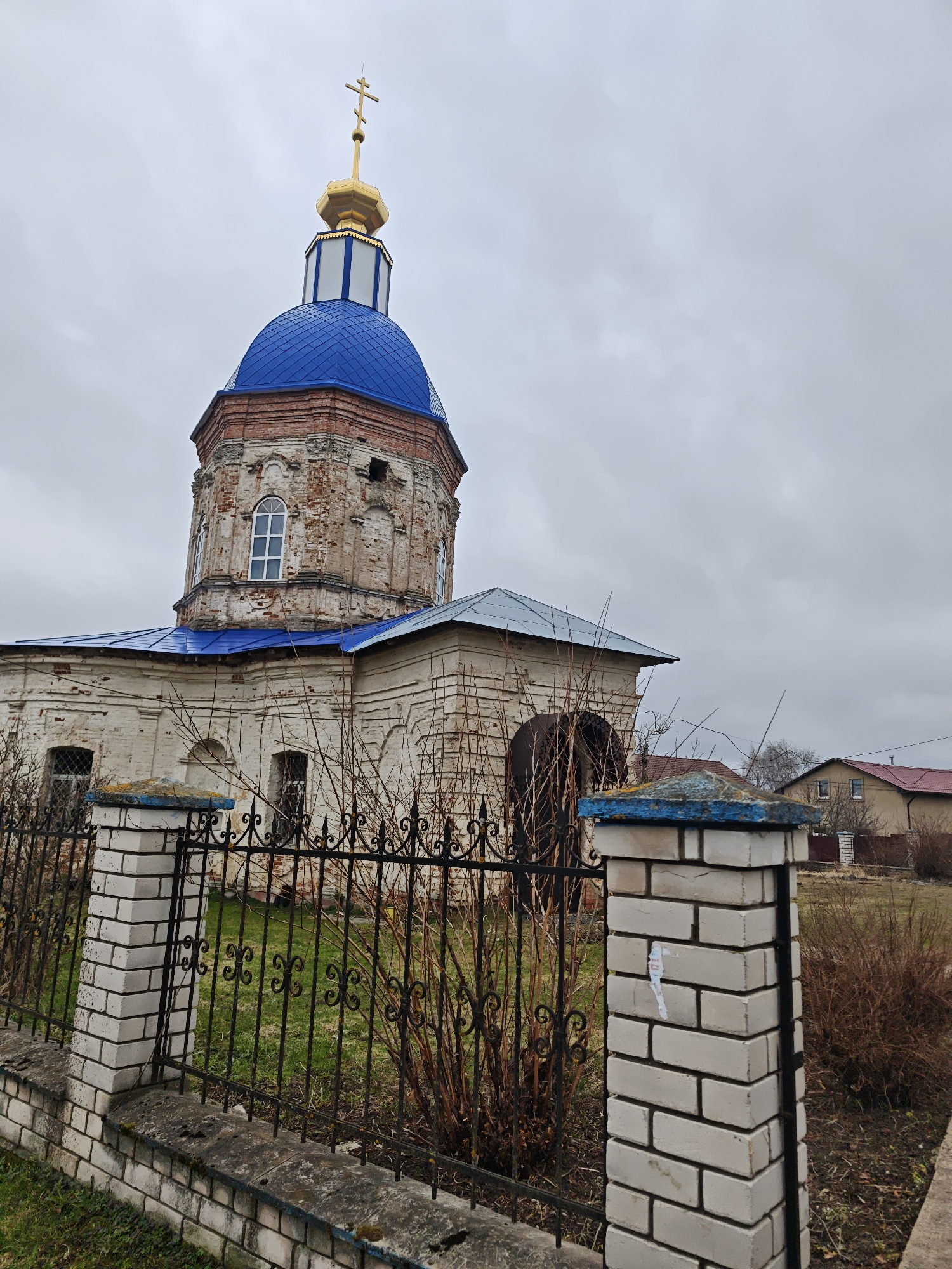 The image shows an old, weathered church in посёлок совхоза "Буденновец", Russia. The church has a blue dome and a golden cross. The walls of the church are made of white brick, and the building is surrounded by a black iron fence with white brick pillars. There are bushes and grass inside and outside the fence. In the background, there are residential buildings and an overcast sky.