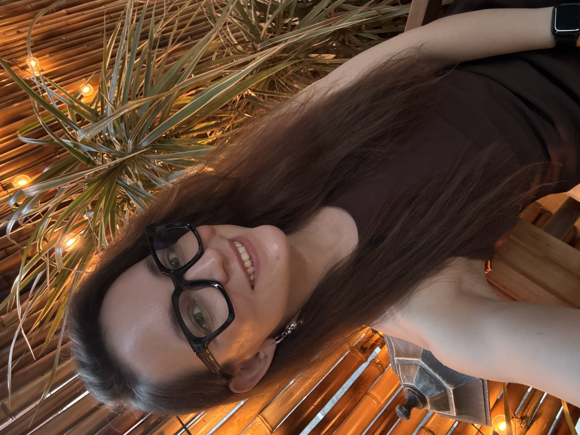 A woman with long brown hair and glasses is posing for a selfie. She is wearing a brown top and a smartwatch. The background includes a plant with long, thin leaves and a bamboo-like structure with lights. There is no indication of the location being Moscow, Russia, or any other specific location.