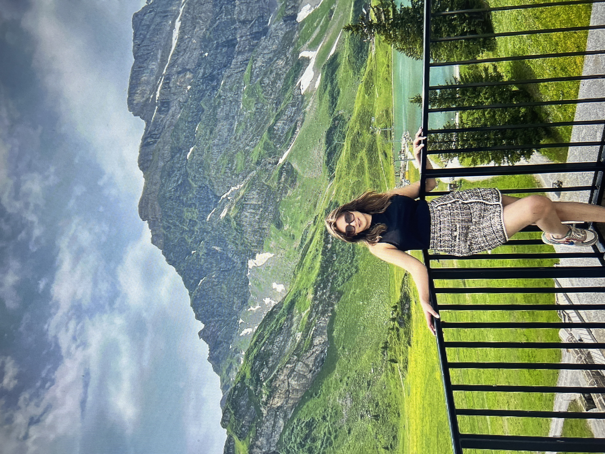 The image shows a young woman standing on a balcony, leaning on a black railing. She is wearing a black sleeveless top, a tweed skirt, sunglasses, and sneakers. The background features a stunning mountain landscape with lush green slopes, patches of snow, a turquoise lake, and a partly cloudy sky. The location is not Qarağandı city administration, Kazakhstan, but appears to be a mountainous region, likely in Europe. The woman is looking at the camera and smiling. The scene conveys a sense of travel and enjoyment of nature.
