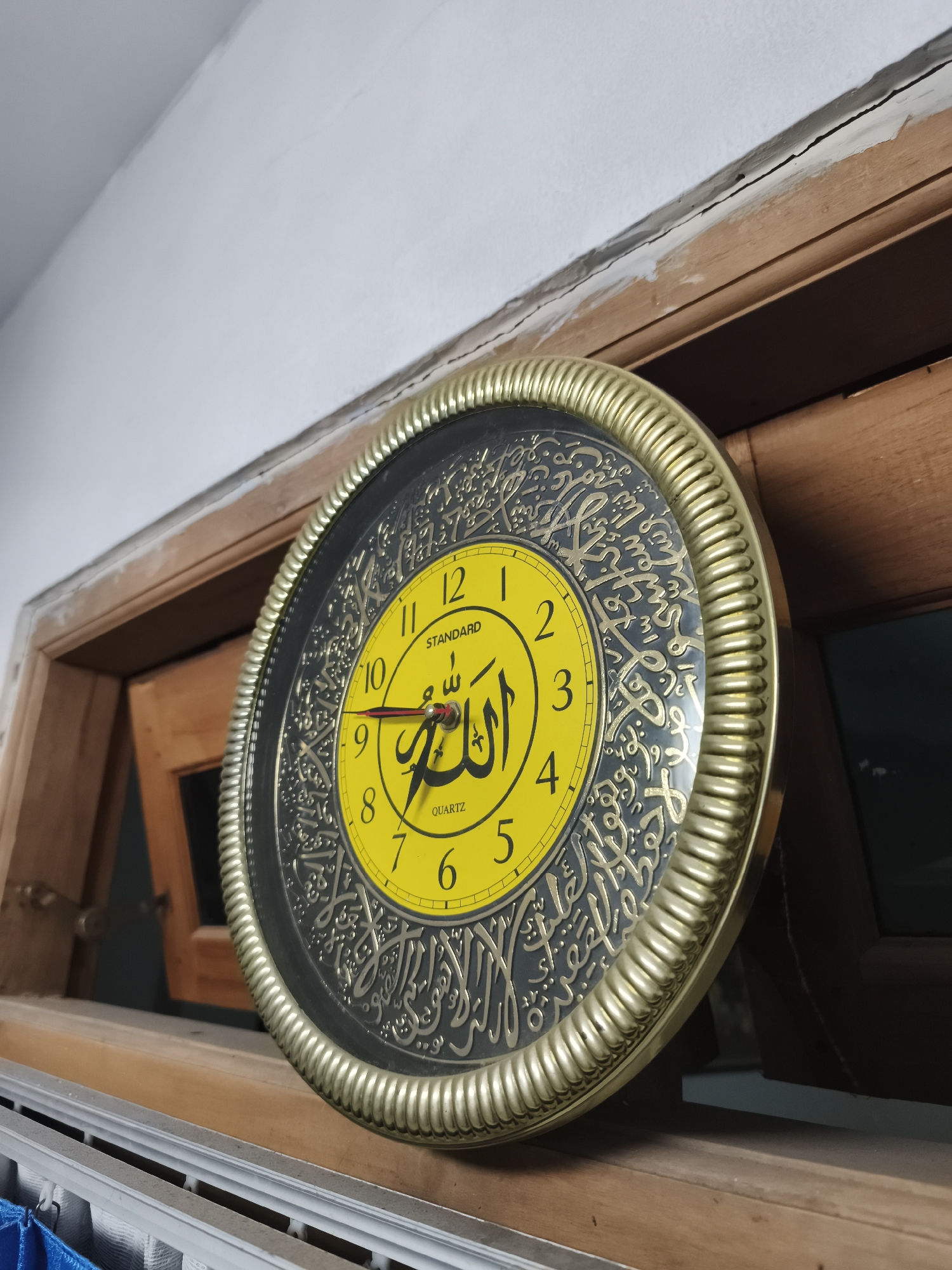 A close-up, eye-level, outdoor shot shows a round, gold-rimmed clock with a yellow face resting on a wooden surface, possibly a window sill. The clock has black Arabic calligraphy design surrounding the yellow face, which displays Arabic numerals and the words "STANDARD" and "QUARTZ." A red hour hand points to the numeral 10, and a black minute hand points slightly past the numeral 9. The clock is positioned against a wooden frame and a white wall. The scene is likely located within the city of Yogyakarta, Indonesia. The image is well-lit, with natural light casting shadows. There are no people or animals visible in the frame.