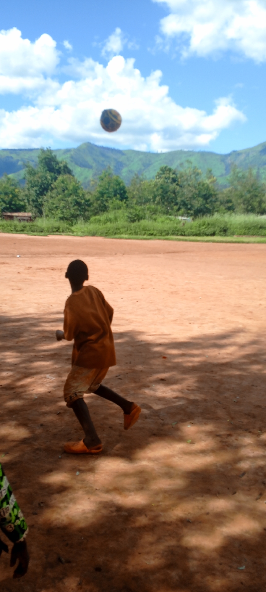 The image captures an outdoor scene in Ngalula, Democratic Republic of the Congo. The sky is bright blue with scattered white cumulus clouds. In the background, a range of green, tree-covered mountains is visible, separated from the foreground by a line of dense green foliage. The ground across the midground and foreground is composed of reddish-brown dirt, exhibiting visible shadows, suggesting direct sunlight.
A primary subject, observed from behind and slightly to the left, is positioned centrally in the midground. This individual wears a loose-fitting, light brown long-sleeved shirt, lighter-colored shorts, and bright orange clogs. The person is captured in mid-stride, with their left leg bent and right leg extended, moving towards the right side of the frame. Their head is turned slightly left, oriented towards a dark, patterned spherical object, identified as a ball, which is suspended in the air above and to the left of the subject. A partial view of a second individual, wearing a green, black, and white patterned garment, is visible in the lower-left foreground. The scene depicts active play involving the ball and at least two individuals.