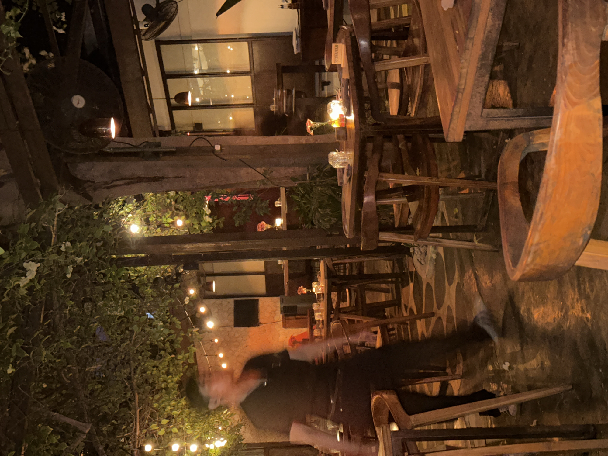 The image depicts an outdoor seating area of a restaurant at night, likely in Kuta Selatan, Indonesia, with an ambiance of string lights and candles. In the foreground, a person in a dark shirt and pants is blurred and in motion, walking past a wooden table set for dining. The table has glasses and a small flower arrangement with a lit candle, suggesting it is ready for customers. The background shows more wooden tables and chairs, with leafy branches of trees overhead. Industrial-style fans are mounted on beams above. The flooring is a textured, dark surface, possibly concrete or stone, with circular patterns visible from lighting. The overall scene conveys a warm and inviting evening atmosphere, common in popular tourist areas of Kuta Selatan.