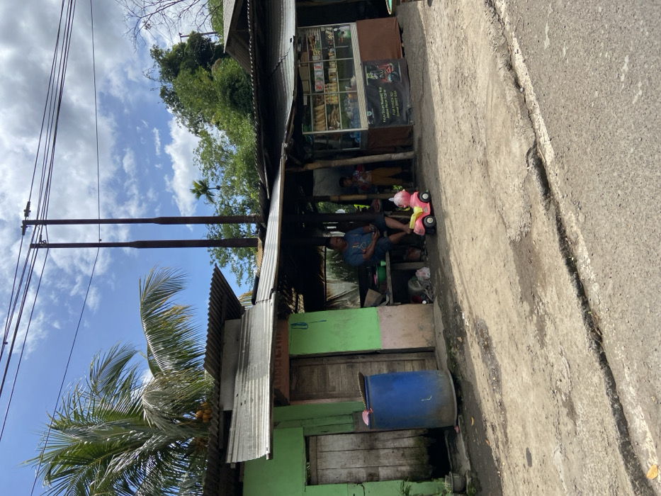 The scene is in Cipatat, Indonesia (latitude: -6.777, longitude: 107.437) and shows a small shop with a corrugated metal roof. There are two men sitting outside, one wearing a blue shirt and the other a red shirt, with a child sitting on a small, pink toy truck. The shop is open and shelves with various goods are visible through the windows. There is a blue barrel in front of the shop. The shop front is adorned with a green sign that reads "Toko" in cursive. The scene is in front of a building that is partially obscured by lush vegetation. There are power lines overhead, and a road in front of the shop. The sky is partly cloudy. The environment is rural and the ground is dirt.