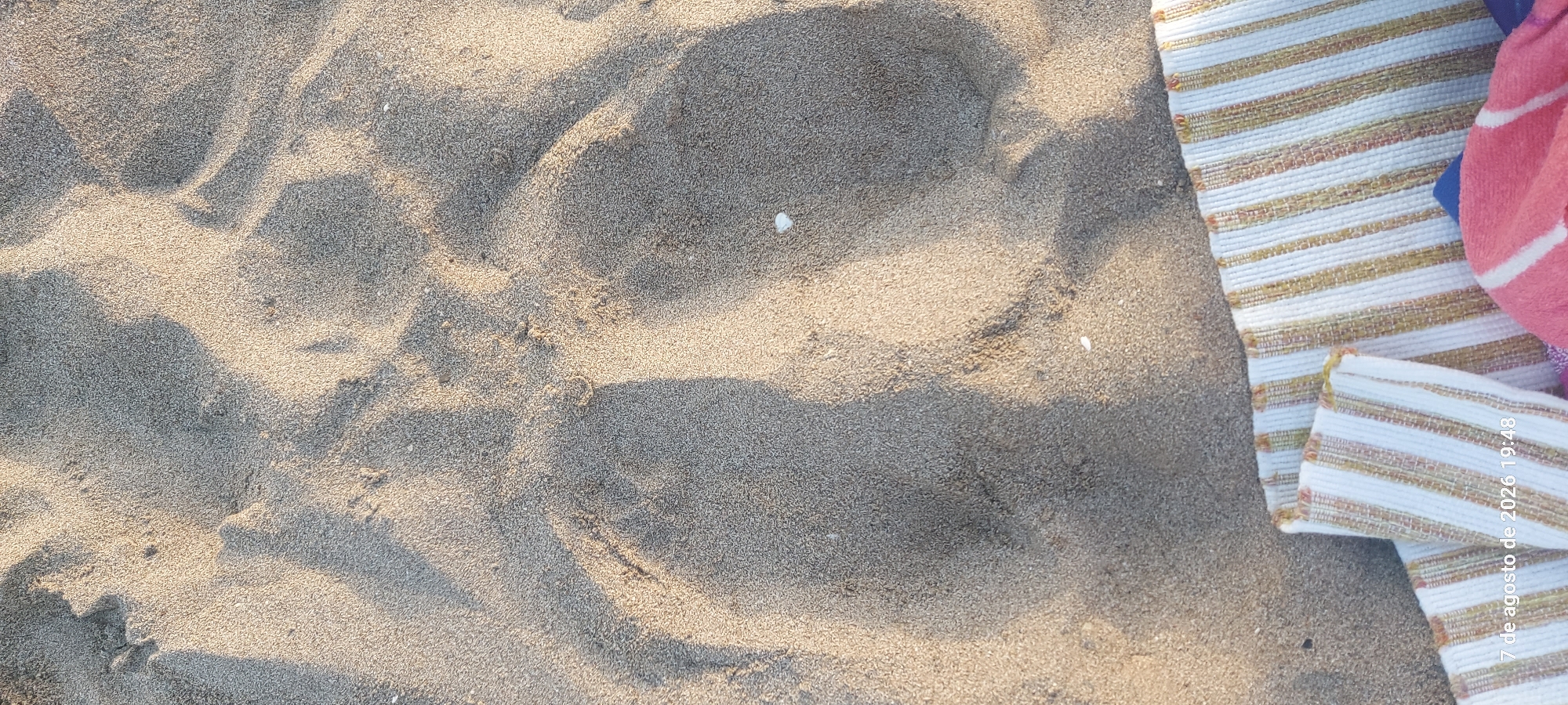 This image shows a close-up view of sand on a beach, with two distinct indentations that appear to be footprints. The sand is light brown and appears to be dry. There are shadows cast across the sand, indicating sunlight from the side. In the lower right corner of the image, a portion of a striped beach towel is visible, along with a hint of a pink towel. The text at the bottom of the image, "7 de agosto de 2026 19:48," indicates the date and time the photo was taken. While the image itself does not provide definitive clues to the exact location within Castelldefels, Spain, the presence of a beach towel and sand suggests an outdoor setting at the coast.
