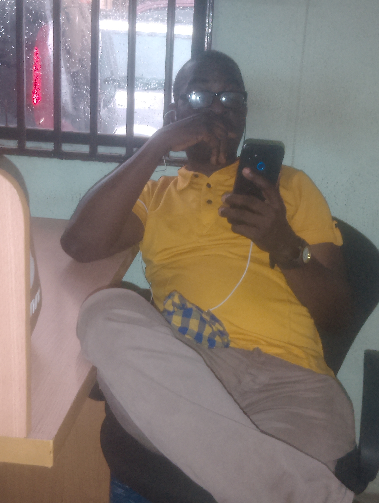 A dark-skinned man wearing glasses, a yellow polo shirt, and light-colored pants is seated and holding a smartphone. He is looking at the phone, which is displaying a blue icon. A checkered cloth is visible on his lap. The man has a watch on his left wrist. He is seated next to a light-colored desk. In the background, a window with bars is visible, and through it, water droplets and a red light can be seen. The scene appears to be indoors.