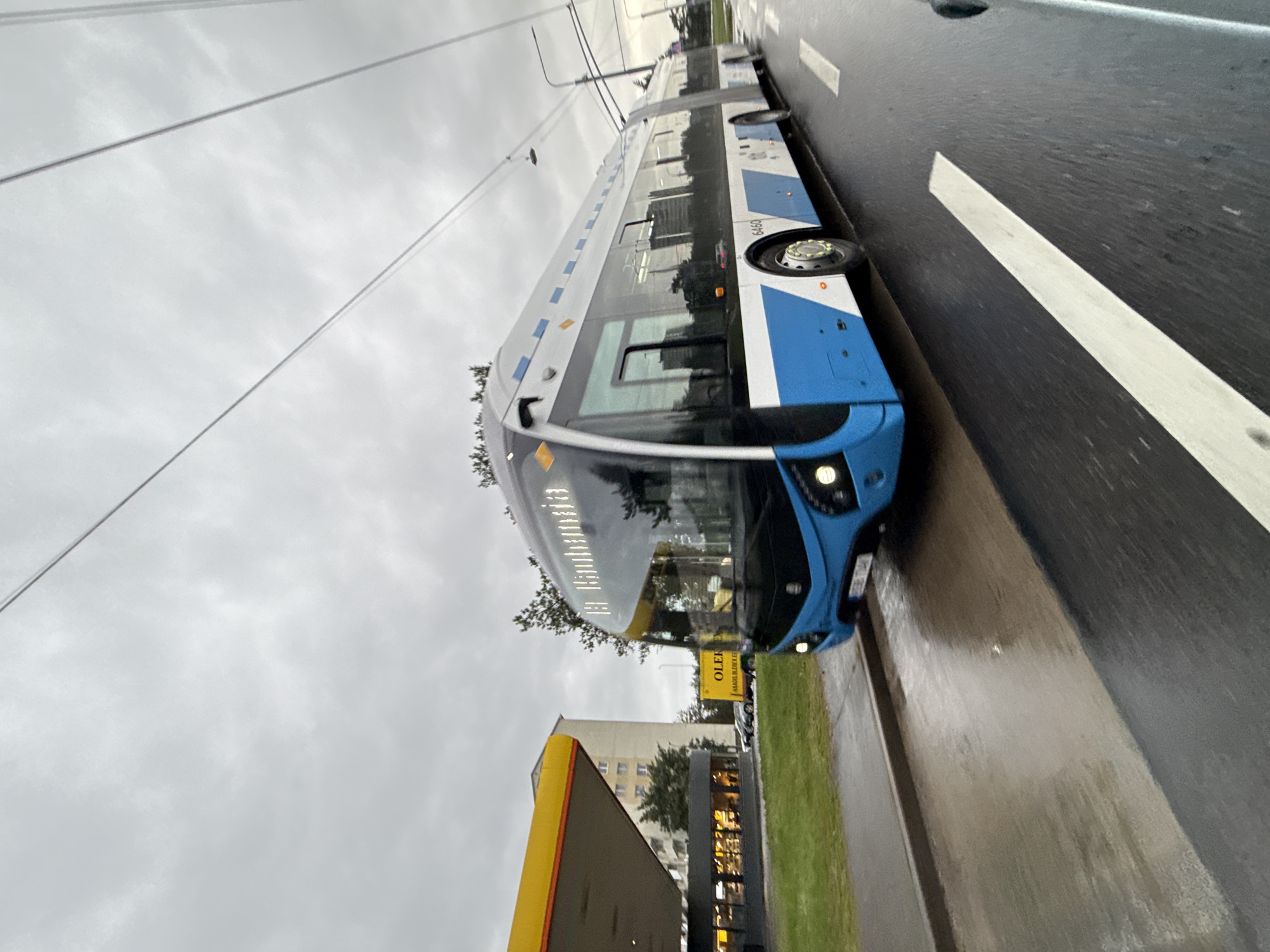 A blue and white articulated trolleybus, identified by the number 6460 on its side, travels on a wet asphalt road under a cloudy sky. The trolleybus is illuminated by its headlights, and its destination sign displays "Pirita". To the left of the road, a yellow and grey structure, likely a gas station canopy, is visible, with a building behind it. In the background, a yellow truck with the text "OLER" is parked. Trees and street lights are also present in the environment. The scene appears to be in Tallinn, Estonia, given the presence of the trolleybus and the likely location of the gas station and building.