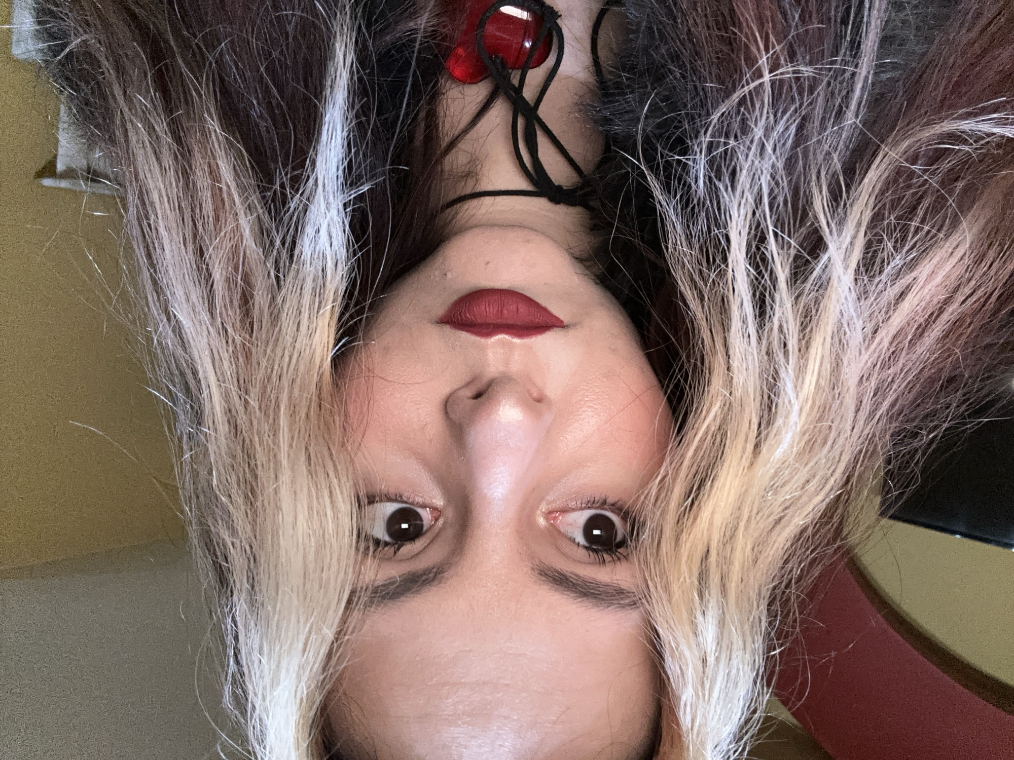 A close-up, upside-down selfie shows a fair-skinned person with brown eyes and dark red lipstick. The person is wearing a black cord necklace with a red stone pendant. The hair is a mix of brown, black, and bleached blonde. The background consists of a yellow wall and part of a red and black chair. The image does not provide information to determine its location in Sofia, Bulgaria, or any specific actions, interactions, or environment.