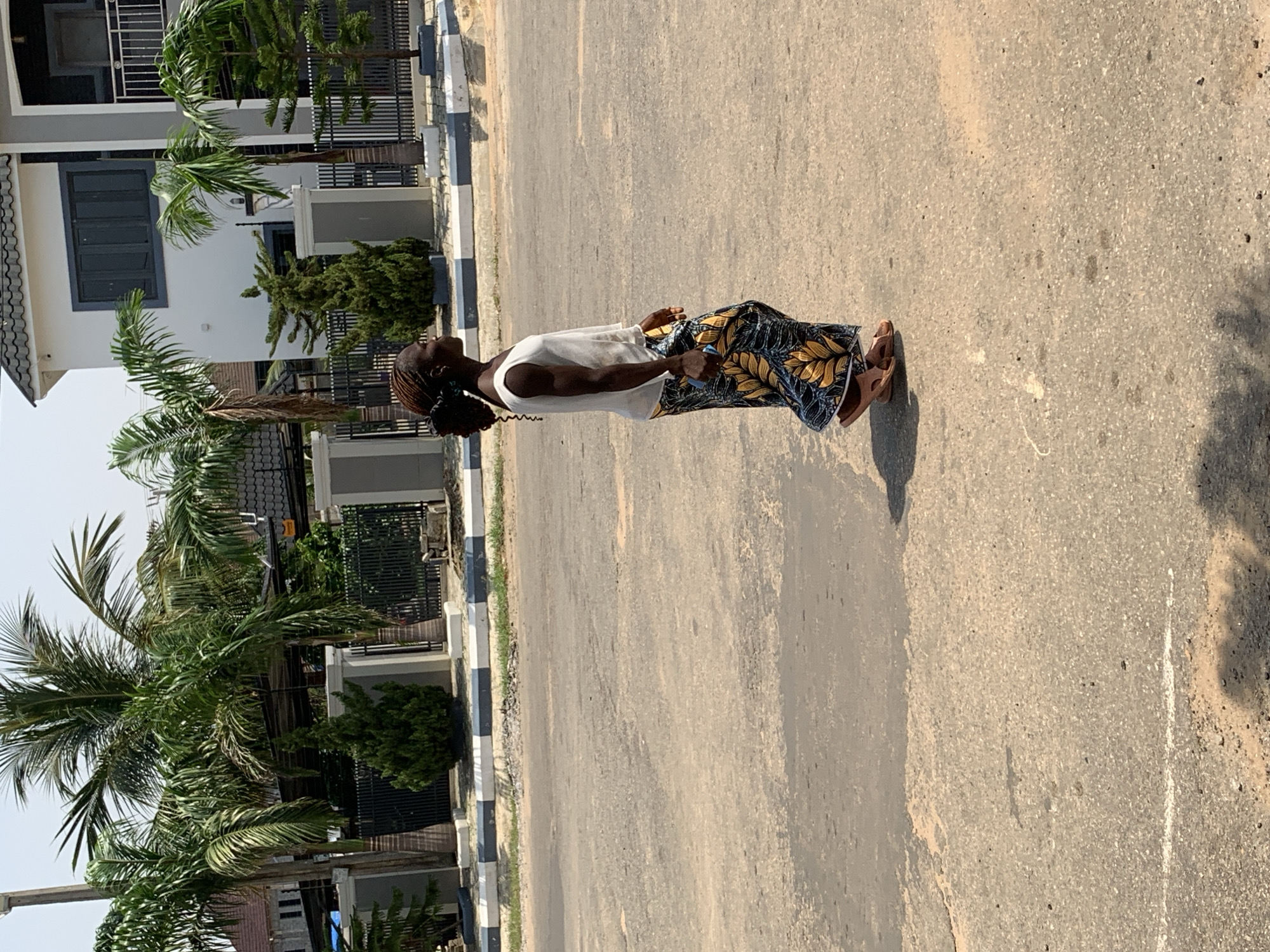 A young Black woman is standing on a paved road in Abuja, Nigeria. She is wearing a white t-shirt and a patterned skirt with yellow and blue designs. The woman has her hair braided in a ponytail. She appears to be looking up and to her right, with a slight smile. In her right hand, she holds what appears to be a small yellow object, possibly a piece of food. Her left hand is by her side. She is wearing brown sandals.
The scene is outdoors, with a clear, bright sky suggesting it is daytime, likely midday due to the strong shadows. The road is asphalt and appears somewhat worn. To the left of the road, there is a sidewalk with a decorative border of black and white stripes. Behind the sidewalk are modern-looking residential buildings with large windows and manicured palm trees and other greenery. The architecture suggests a relatively affluent neighborhood. The overall impression is one of a casual, everyday moment captured on a sunny day. No visible text is present in the image.