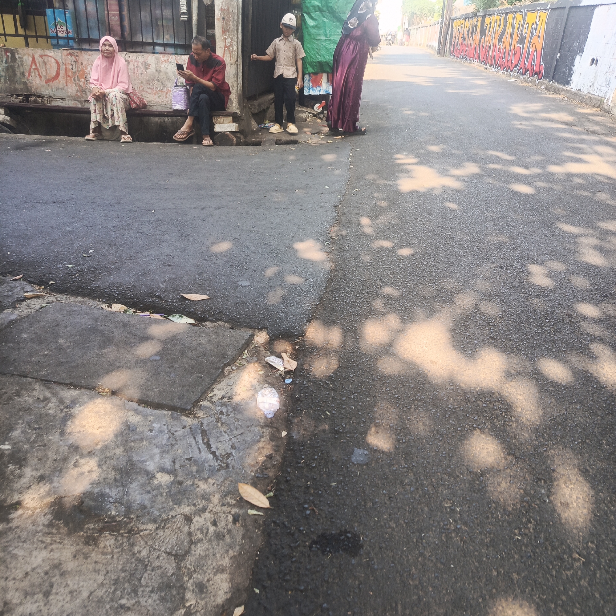 The image depicts a street scene in South Jakarta, Indonesia, characterized by an asphalt road, a sidewalk, and a building facade. In the foreground, a sidewalk area shows some debris including leaves and plastic. The asphalt road surface is partially shaded by dappled light, suggesting trees or structures overhead.
On the sidewalk, a woman wearing a pink hijab and a patterned skirt is seated on a bench. To her right, a man in a maroon shirt and dark pants is also seated, looking at his mobile phone. Further to the right, a young boy in a light-colored shirt and a white cap stands near a green tarp. Beside him, a person in a dark purple hijab and dress is standing. The building facade behind them is weathered and features graffiti.
The overall environment suggests an urban setting, likely a residential or commercial area, with everyday activities taking place outdoors. The presence of a bench, people interacting with their phones, and street art implies a community space or a transitional area.