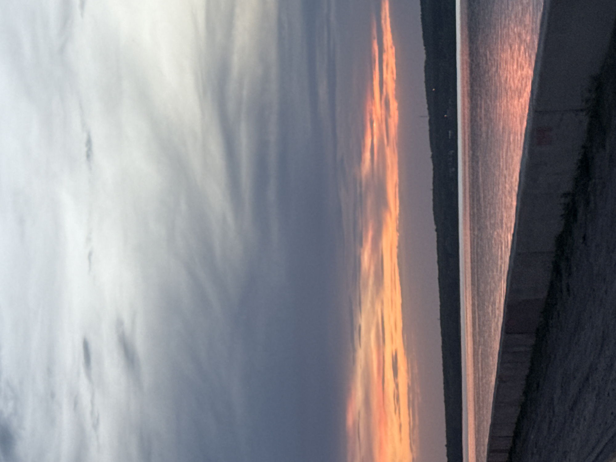 The image shows a view of the Kama River in Chaikovsky, Russia, at sunset. The sky is partially cloudy, with the setting sun casting orange and pink light on the clouds. The river surface reflects the colors of the sunset. A concrete embankment lines the riverbank in the foreground, with some vegetation present. Across the river, a dark tree-covered shoreline is visible.