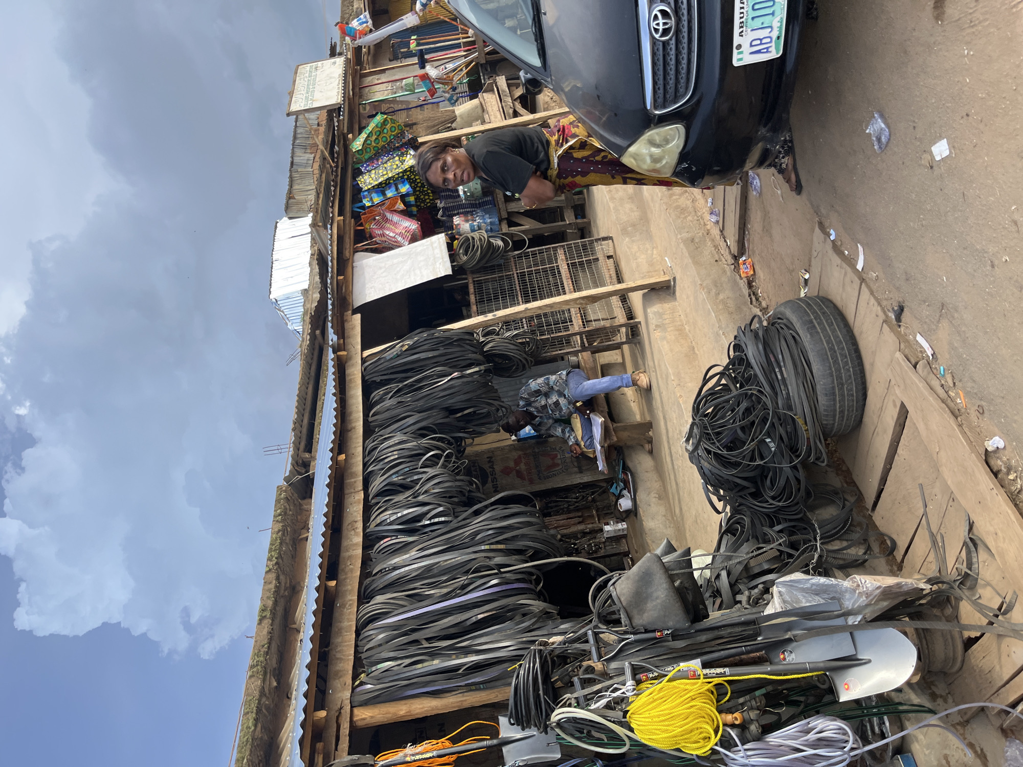 In Rafin Par, Nigeria, a street market bustles with activity. A woman in a colorful patterned skirt and dark t-shirt stands near a black car with a license plate reading "ABJ-102". She looks towards the camera, her expression neutral. The car is parked in front of a stall overflowing with black rubber belts, coiled and piled high. These belts, some appearing to be for vehicles, are the primary merchandise on display.
Behind the woman, the market stall is a colorful assortment of goods. Bags in vibrant patterns are stacked, and various items are displayed on shelves. A man in a patterned shirt and blue jeans is visible further back, seemingly engaged in work at another stall. The scene is outdoors, with a cloudy sky overhead, suggesting it could be any time of day. The ground is dusty and uneven, typical of a market in a developing area. The overall impression is one of a busy, active marketplace where goods are being sold and transactions are taking place. The focus is on the variety of rubber belts and the commerce of the market.