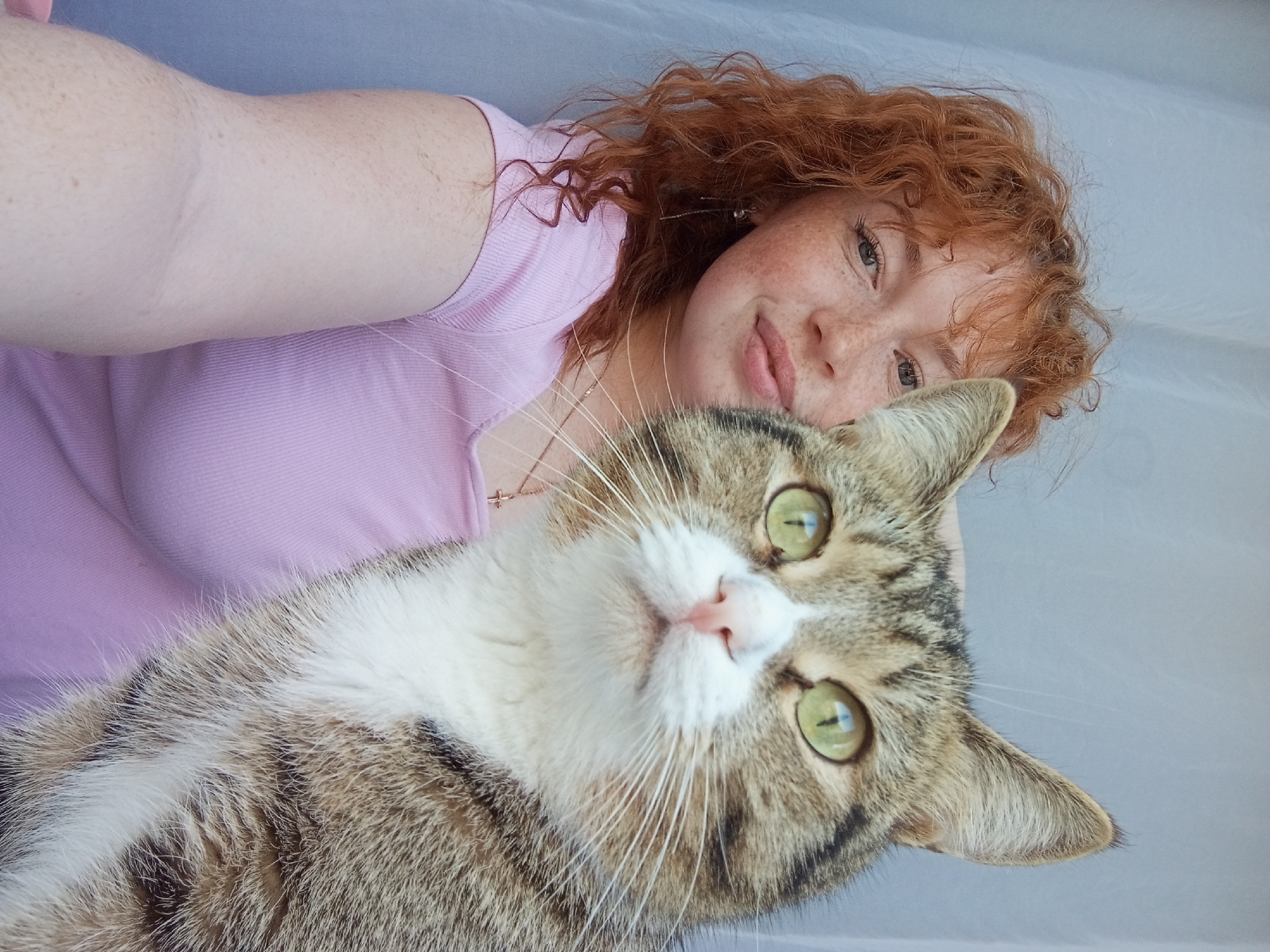 A selfie-style close-up photograph depicts a young woman and a tabby cat. The woman has fair skin, freckles, and red curly hair. She is wearing a pink t-shirt and a gold necklace with a cross pendant is visible. Her head is positioned above the cat's. The cat has green eyes and a mix of brown and white fur. The background appears to be a light-colored, possibly white, sheet or wall. The location is not discernible from the image. The picture suggests a close relationship between the woman and the cat.