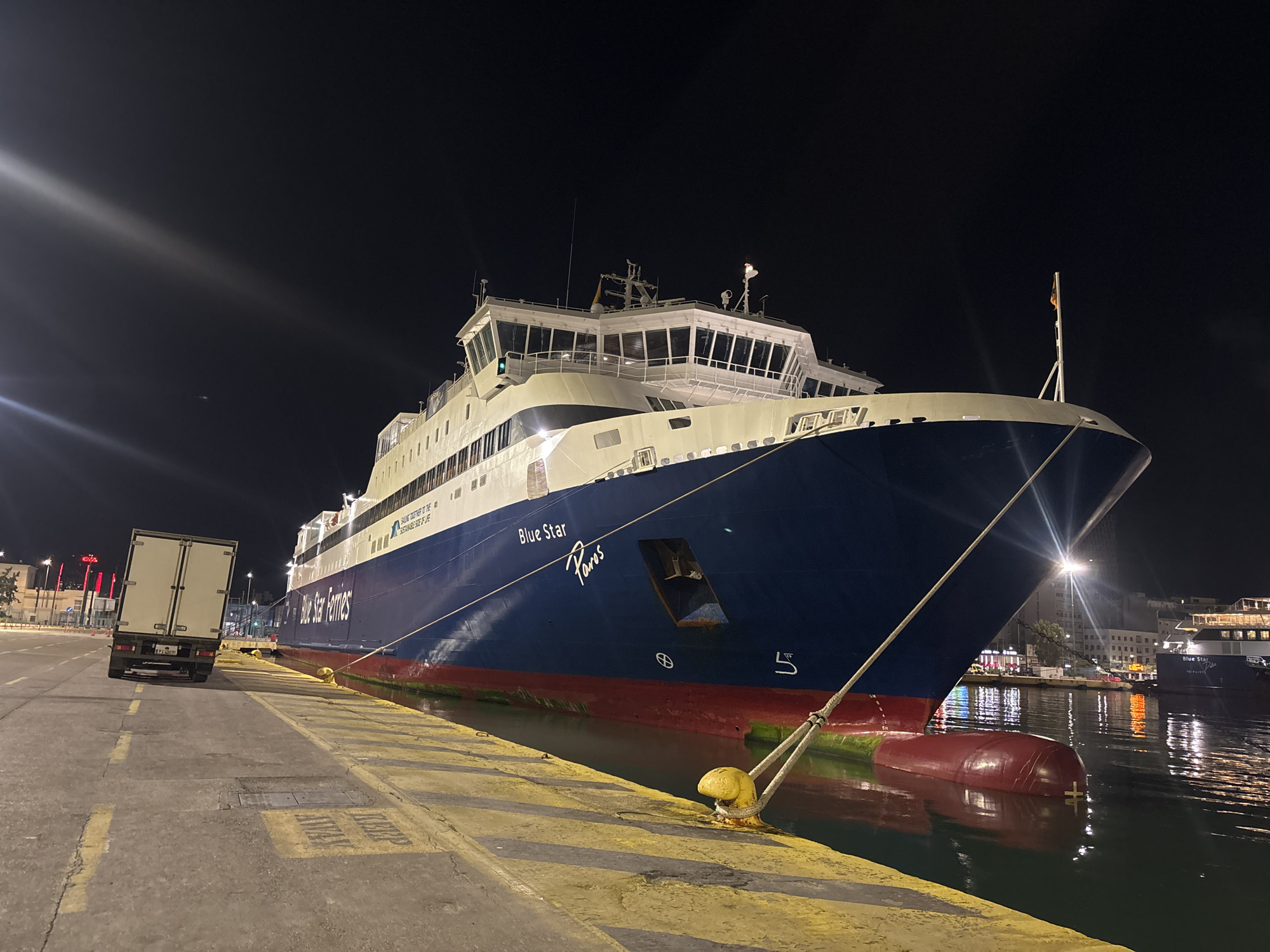 At night in Piraeus, Greece, a Blue Star Ferries vessel named "Paros" is docked. The ship, painted in navy blue, red, and white, is secured to a concrete pier with yellow bollards. A white box truck sits parked on the pier next to the vessel. In the background, lights from the city reflect on the water. Another smaller vessel is docked nearby.