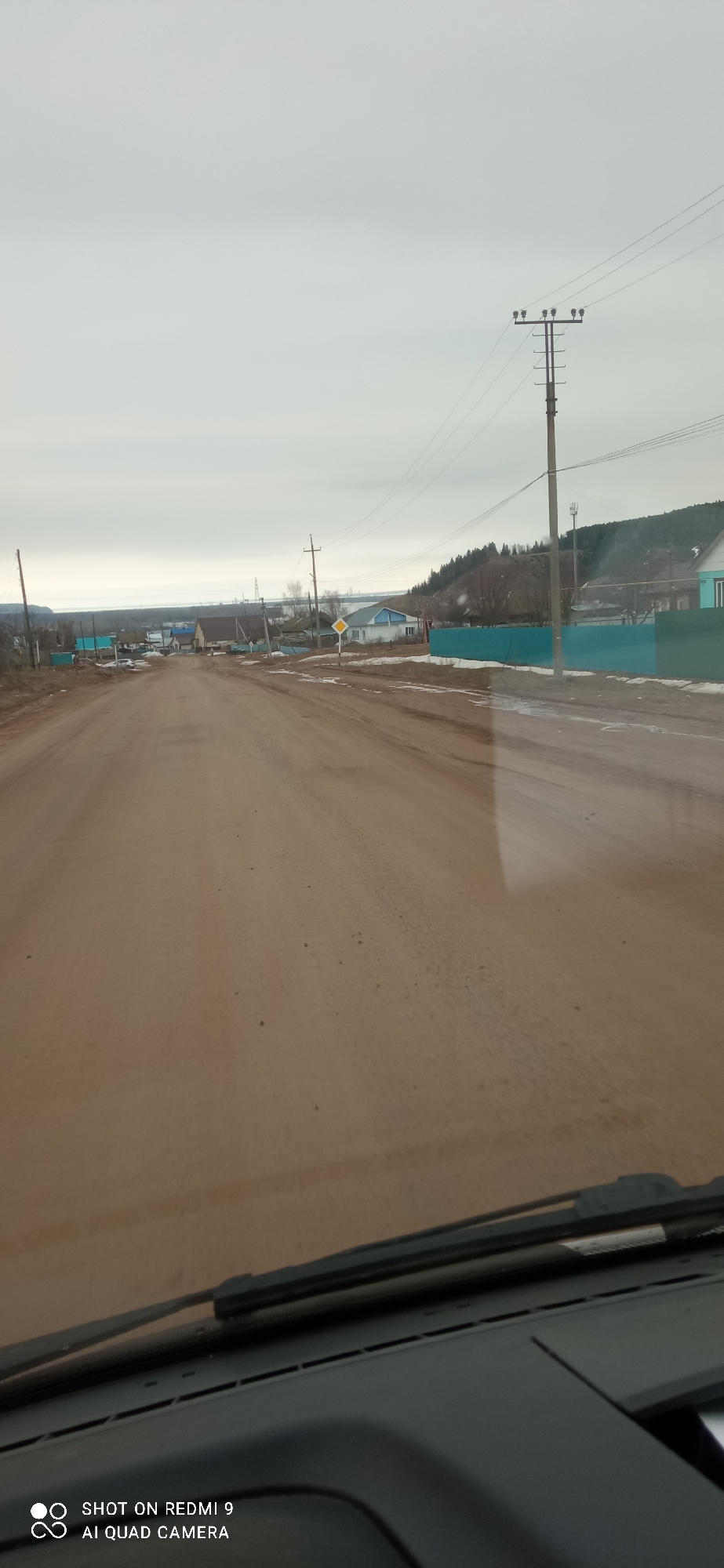 The image depicts a dirt road in the rural area of Ольховка, Russia. The view is from inside a vehicle looking out through the windshield, with part of the dashboard visible. The road is unpaved and muddy, with tire tracks. The sky is overcast. Houses and fences line the road, with utility poles and power lines visible. A small hill is visible in the background. There are patches of melting snow near the fence. The scene shows the typical environment of a village, with a basic infrastructure.