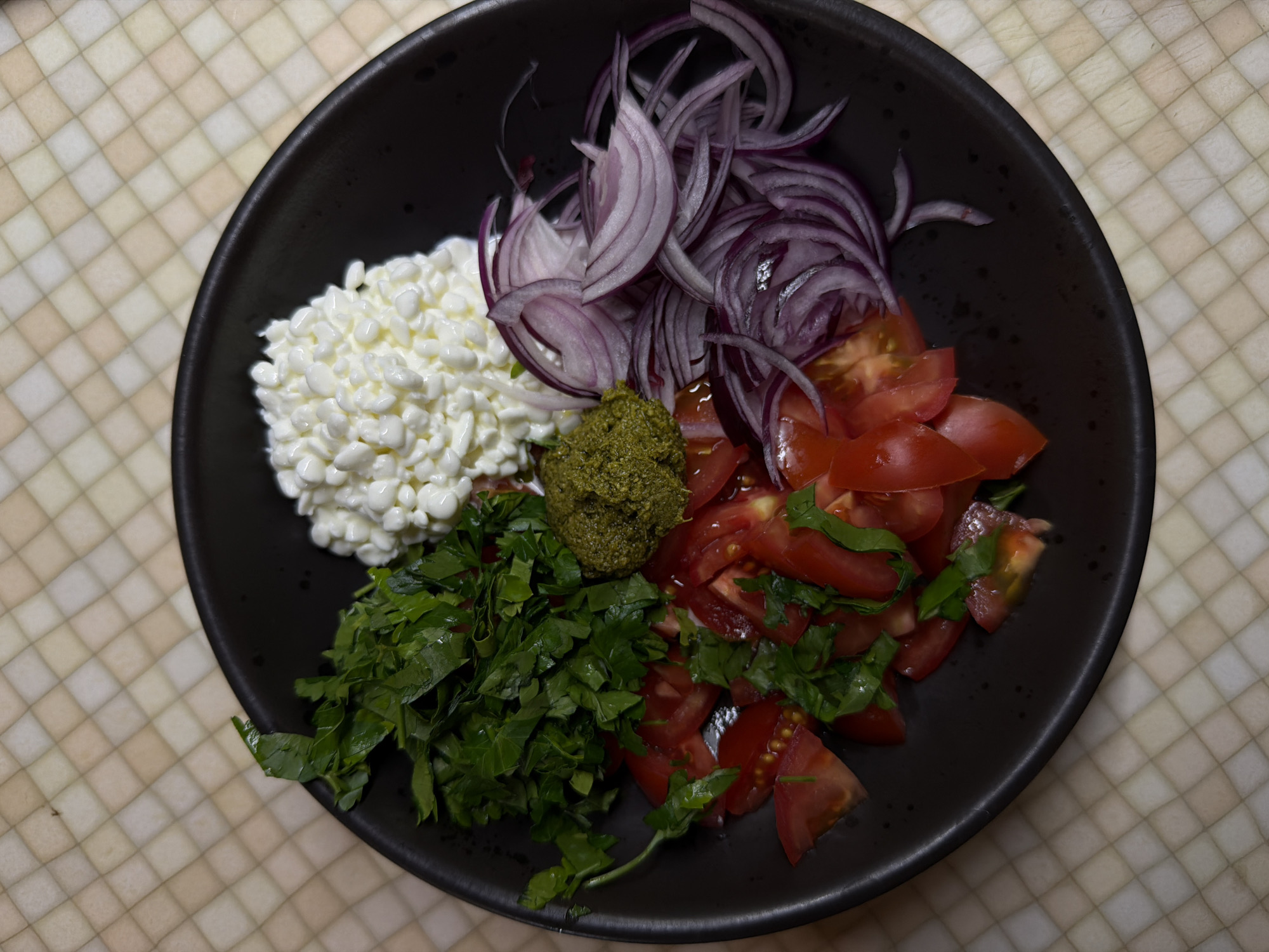 A black bowl is filled with a composition of diverse food items, set against a tiled background. The bowl contains cottage cheese on the left, thinly sliced red onions towards the top, and chopped tomatoes mixed with herbs in the lower right quadrant. At the center, there is a dollop of what appears to be pesto. No people are visible, and there is no indication of location in Khimki, Russia.