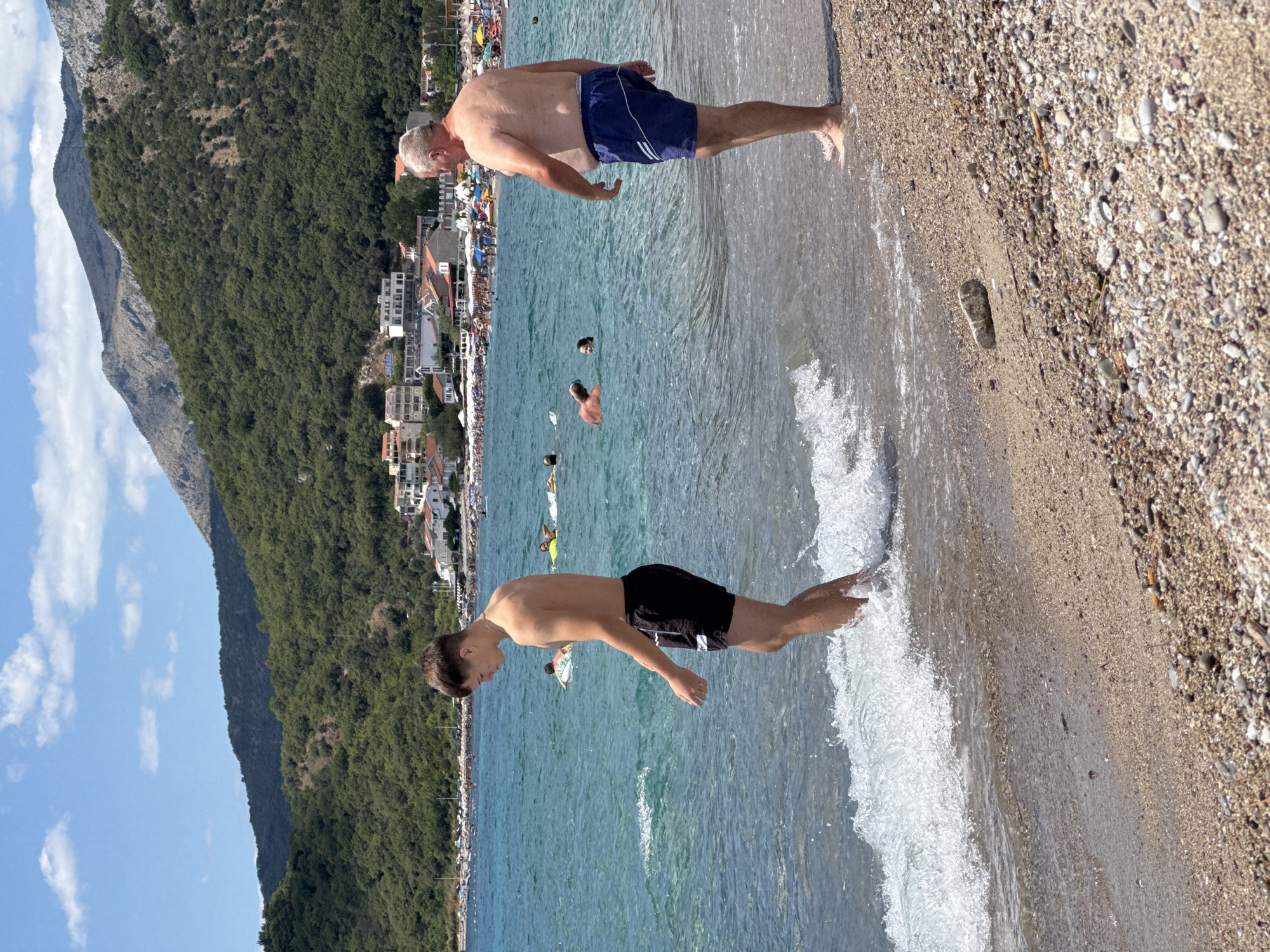 In the city of Đurovići, Montenegro, two men are present on a beach. The younger man, on the left, is standing in shallow water near the shore, facing left and looking down at the waves. He is wearing black swim shorts. The older man, on the right, is standing on the sandy and pebbly shore, facing away from the camera and towards the water. He is wearing dark blue swim shorts. Several other people are visible in the turquoise water in the middle ground, some swimming and others floating on tubes. In the background, a crowded beach with umbrellas and buildings lines the coast, backed by a lush green, tree-covered mountain range under a bright blue sky with scattered clouds. The overall scene depicts a sunny day at the beach.
