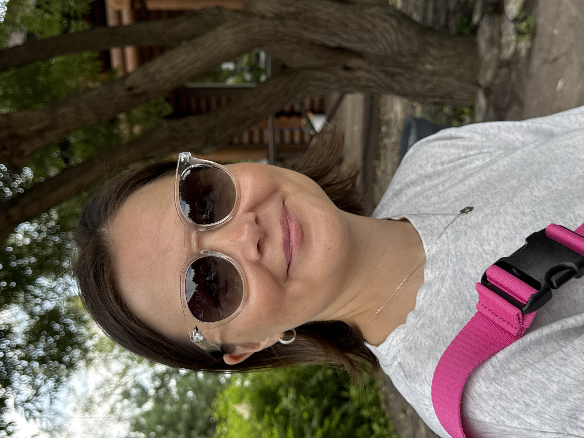 A woman with medium-length brown hair and sunglasses smiles at the camera. She is wearing a light gray t-shirt with a bright pink strap across her shoulder, likely from a bag. She has a silver necklace with a pendant and a hoop earring in her left ear. In the background, trees and a wooden building are visible. The image is taken outdoors, suggesting a park or natural setting within Izhevsk, Russia. The lighting is natural, indicating it is daytime.