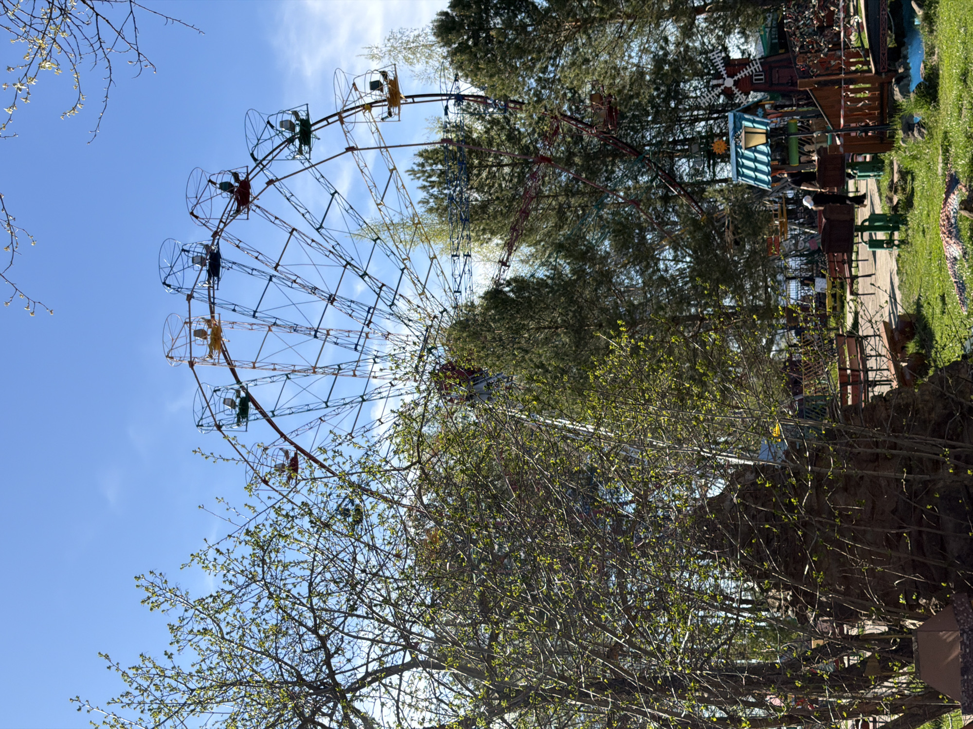 The image shows a Ferris wheel located in the city of Chaikovsky, Russia. The Ferris wheel is made of metal and has colorful gondolas. The image is taken from a low angle, looking up at the Ferris wheel. The sky is blue and there are trees in the background. In the midground are bushes and trees, and an outdoor play area can be seen to the right of the frame.