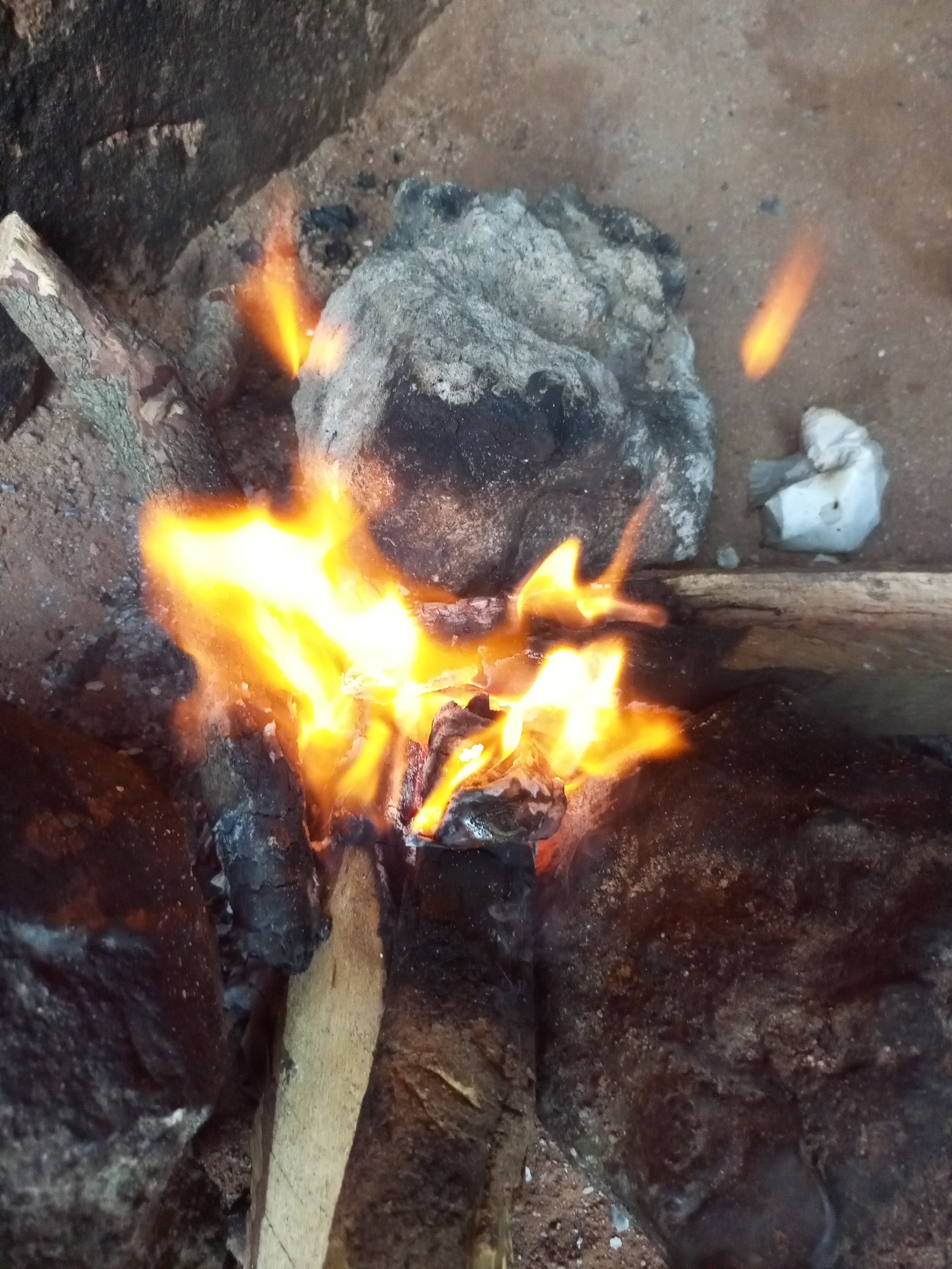 A close-up, overhead view shows a small, open fire burning within a rustic hearth. The fire is fueled by pieces of wood and what appear to be rocks or coals, casting a bright orange and yellow glow. The surrounding environment is composed of earth and rough, textured surfaces, suggesting an outdoor or rudimentary setting. There are no people or animals visible in the image. The location is identified as Tempure, Nigeria.