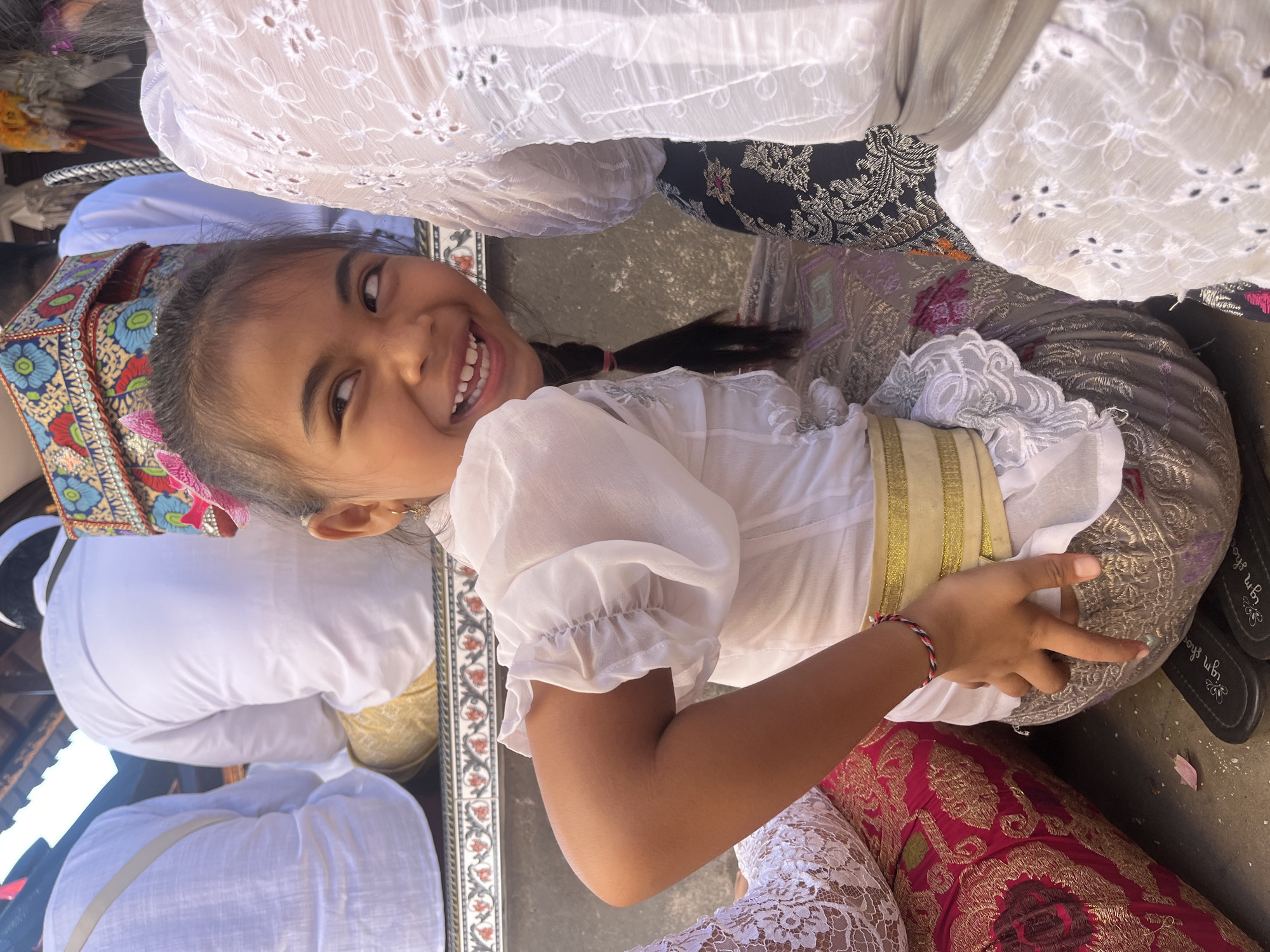 A young girl with dark hair styled in a braid and a pink bow smiles and looks to her right while sitting on the ground. She wears a white blouse with ruffled sleeves and a gold sash. She is surrounded by people in white clothing, and colorful textiles are visible on the ground. The scene appears to be outdoors, possibly during a cultural event or ceremony. Based on the attire and the presence of what looks like traditional Indonesian religious adornments, it is likely this image was taken in Mendoyo, Bali, Indonesia.