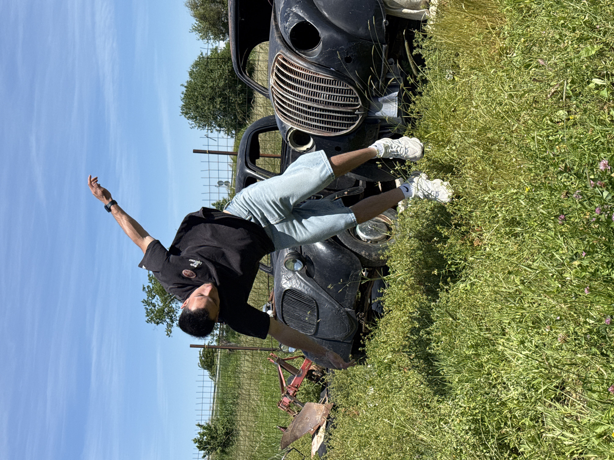 A man in a black t-shirt, light blue shorts, and white sneakers is balancing on his hands in a grassy field. He is positioned between two rusted, black car bodies, one on top of the other. The man's right arm is extended upwards towards a blue sky, while his left arm is grounded near the lower car body. A fence is visible in the background, with sparse trees beyond it. The location is presumably in or near Omurtag, Bulgaria, based on the user request.