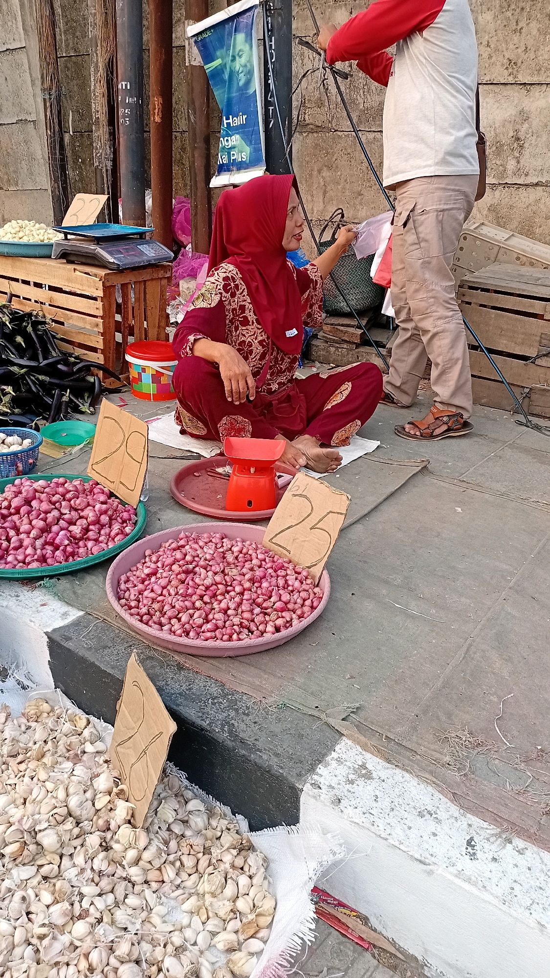 A street vendor in Jambi, Indonesia, sits cross-legged on the ground beside her produce stall. She wears a maroon hijab and a matching patterned garment. In front of her are large trays of shallots and baskets of garlic. A red scale sits on a tray, possibly used for weighing produce. Behind her, a man stands, seemingly engaged in a transaction with the vendor. The market appears to be on a sidewalk, with a stone wall and wooden crates visible in the background. The ground is paved with concrete. A sign with an image of a man and text in Indonesian is attached to a pole.