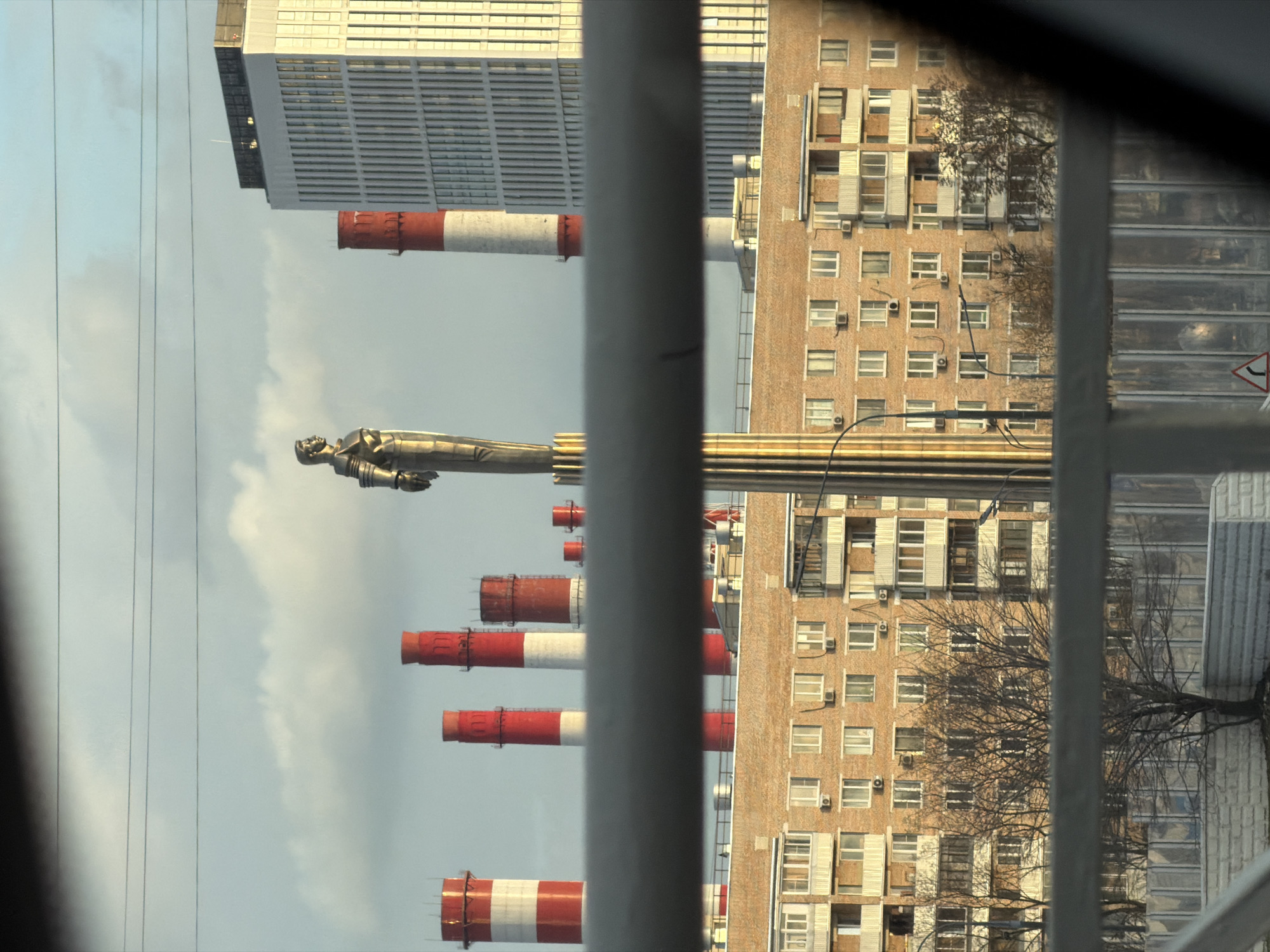 The image shows a view through a window with vertical bars, capturing a scene in Moscow, Russia. In the background is a statue atop a tall column, likely the Monument to the Conquerors of Space. Several industrial chimneys with red and white stripes are visible. Buildings are also present, including a modern high-rise and an older brick building with numerous windows and air conditioning units. Power lines are visible against a sky with some clouds.